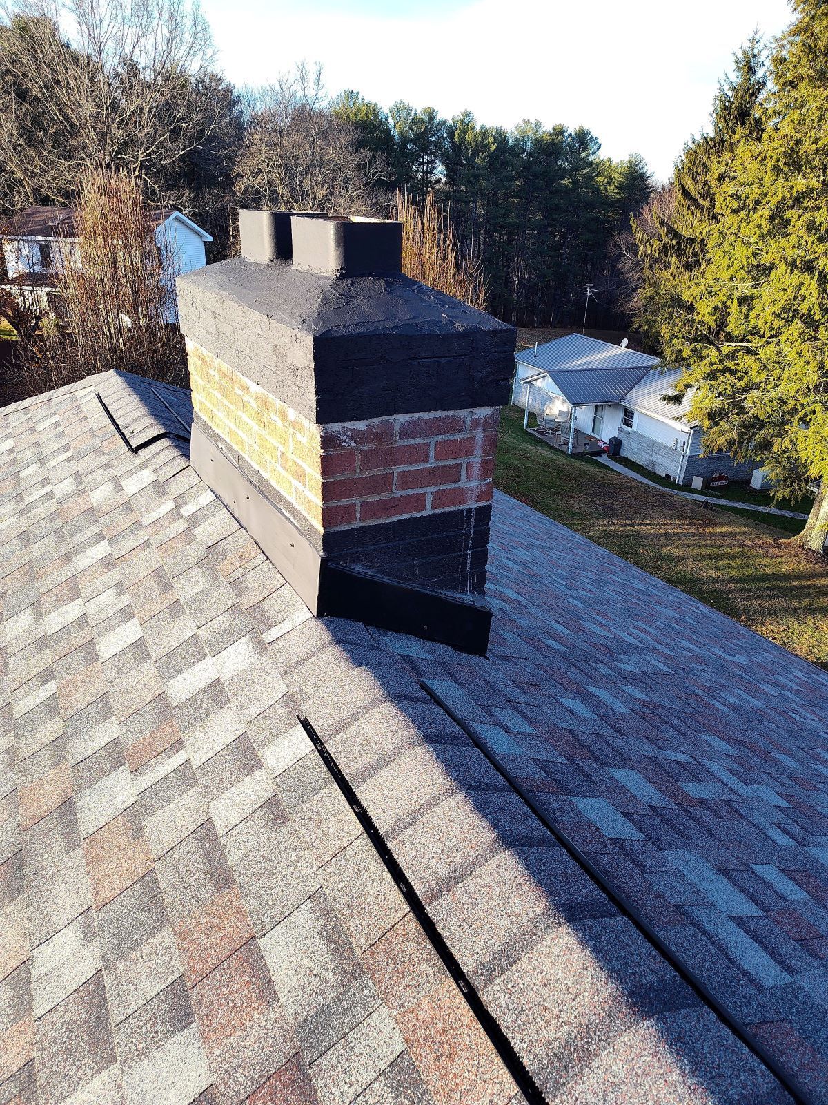 A brick chimney with a black cap and flashing on a shingled residential roof, overlooking a yard and nearby house.