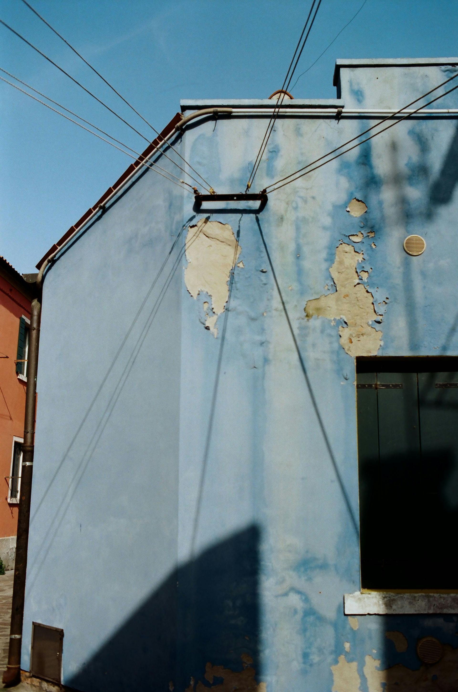 A blue building wall with peeling paint under a bright blue sky, crisscrossed by overhead wires.