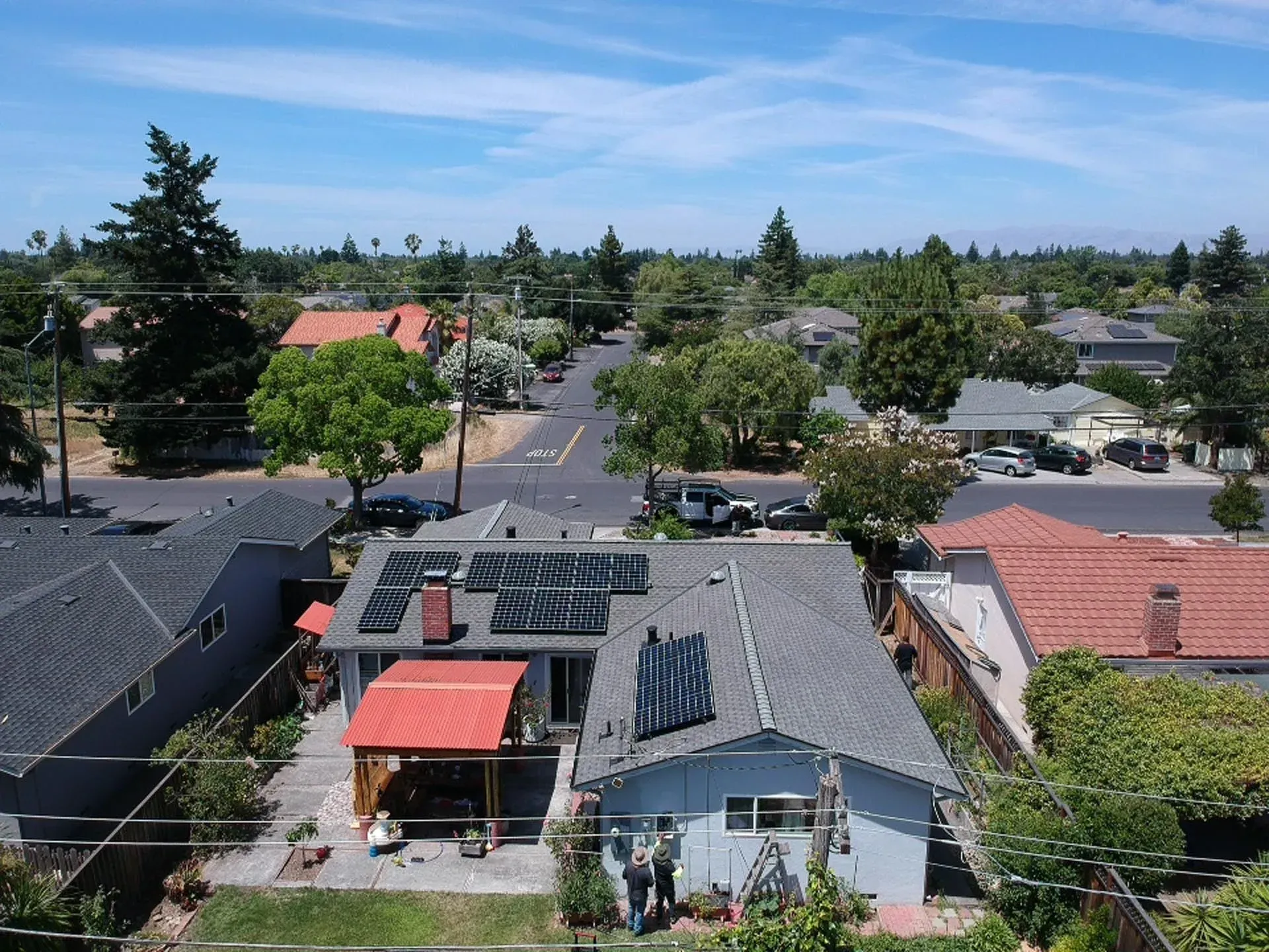 An aerial view of a gray residential house with solar panels on its roof, a red patio canopy, and a suburban neighborhood.