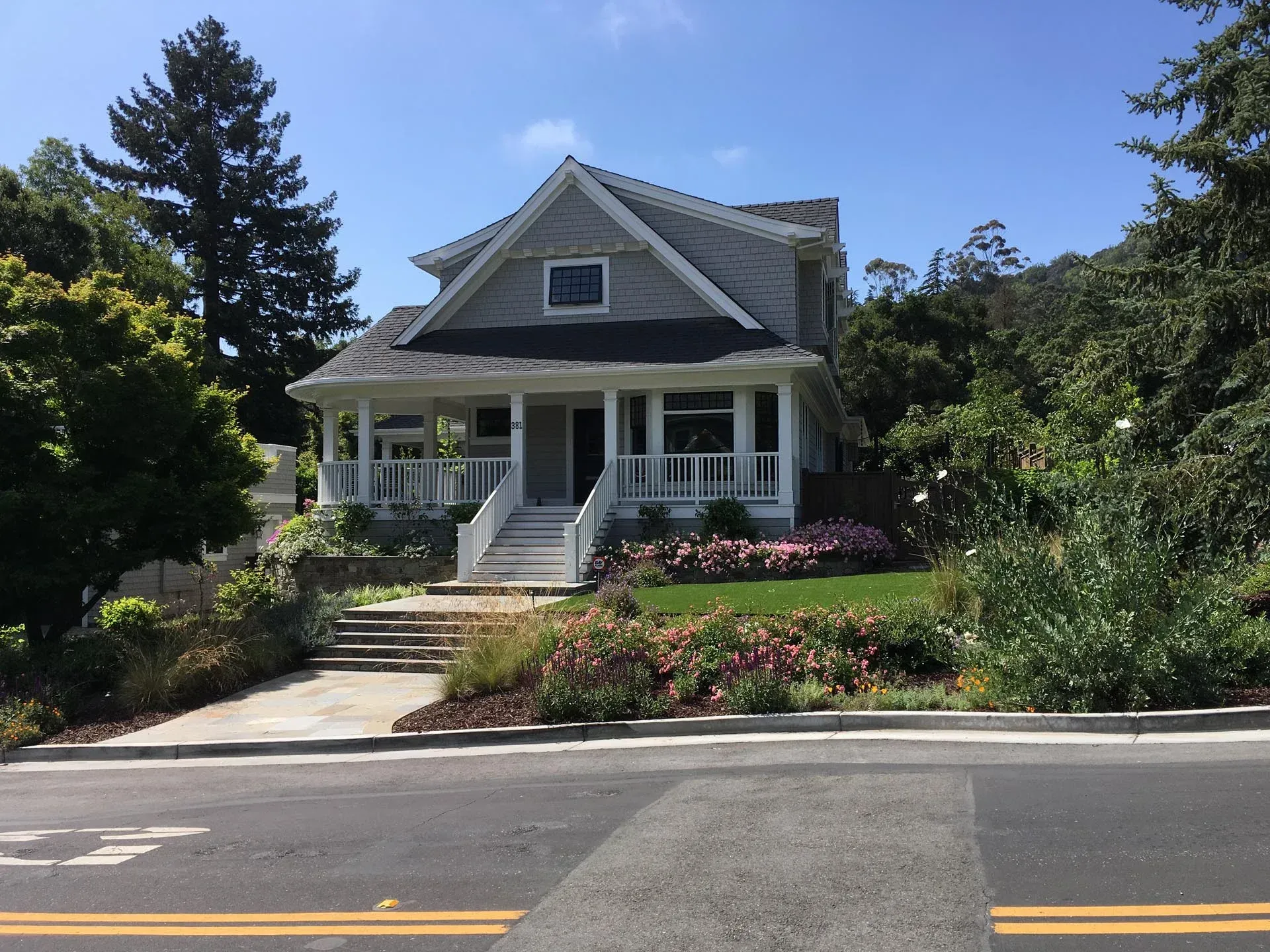 A gray two-story house with a white front porch and steps, set behind a landscaped yard with flowers and large trees.