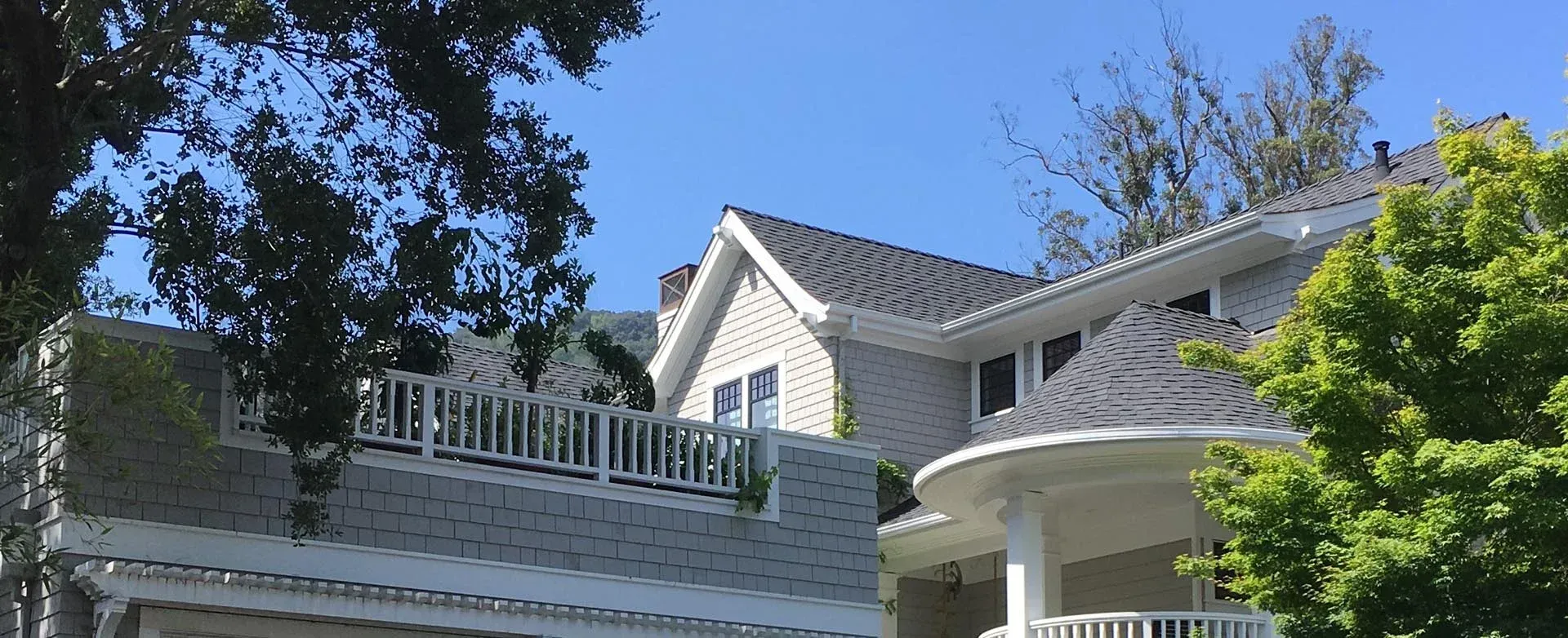 A light-colored house with cedar-shingle siding, a white balcony railing, and a turreted roof under a clear blue sky.
