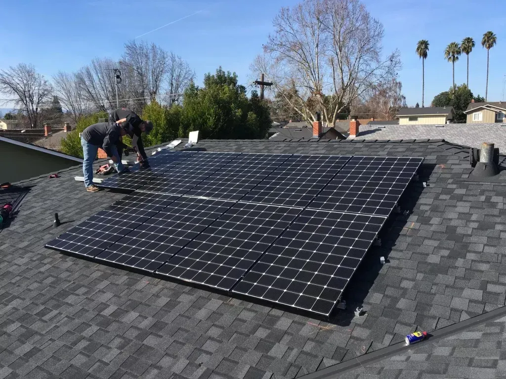 A worker installs a rectangular array of black solar panels on a dark gray shingled roof under a clear blue sky.