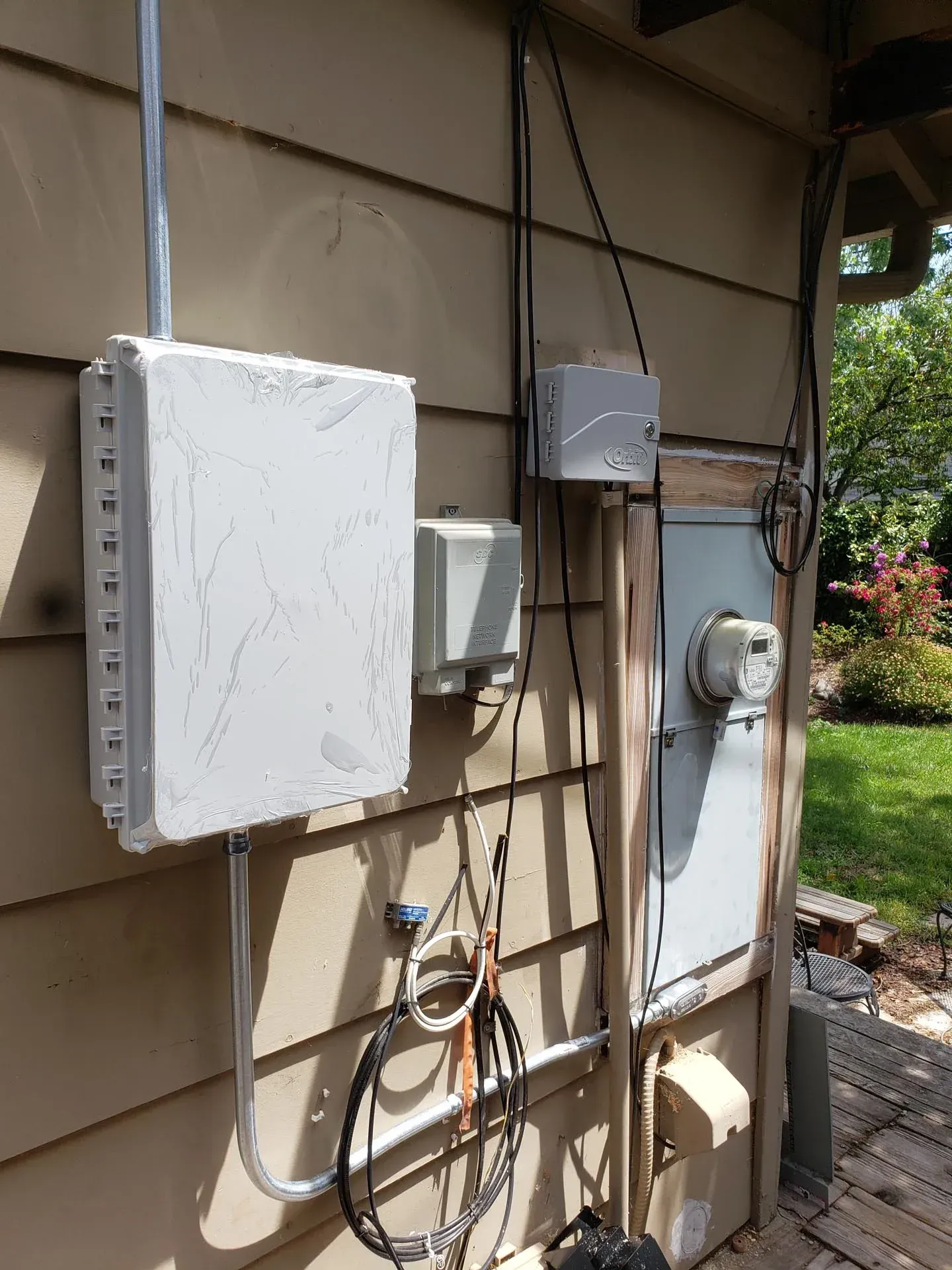 Utility equipment on a wood-sided wall, including a large white electrical box, a utility meter, and various cables.