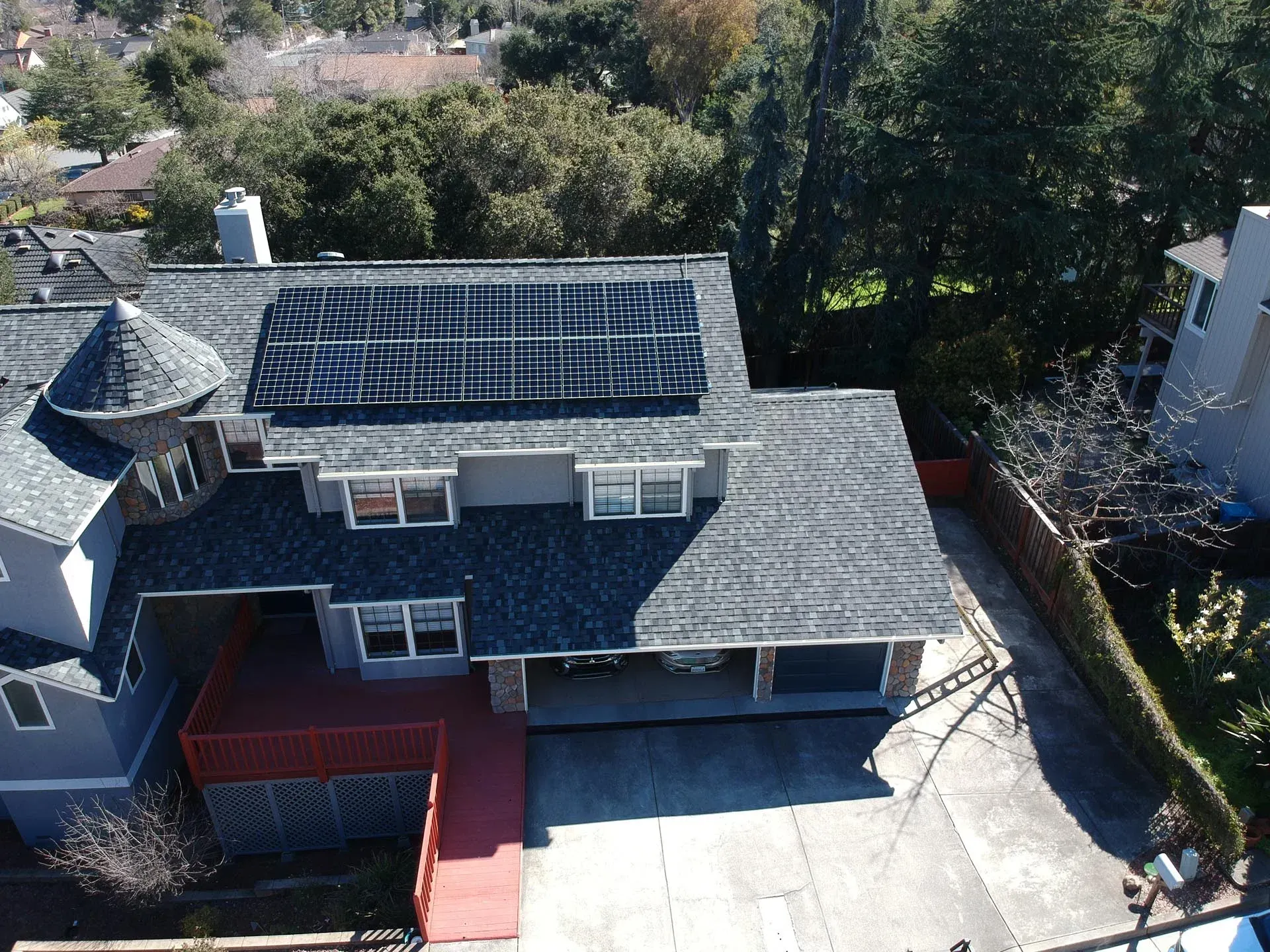 An aerial view of a multi-story, grey-sided house with a shingled roof, solar panels, and a red deck.