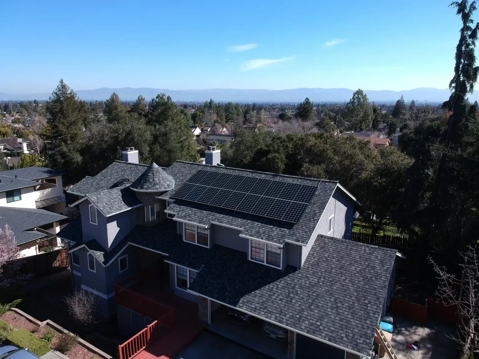 Aerial view of a gray, multi-story house with a shingled roof, solar panels, and surrounding trees under a clear blue sky.