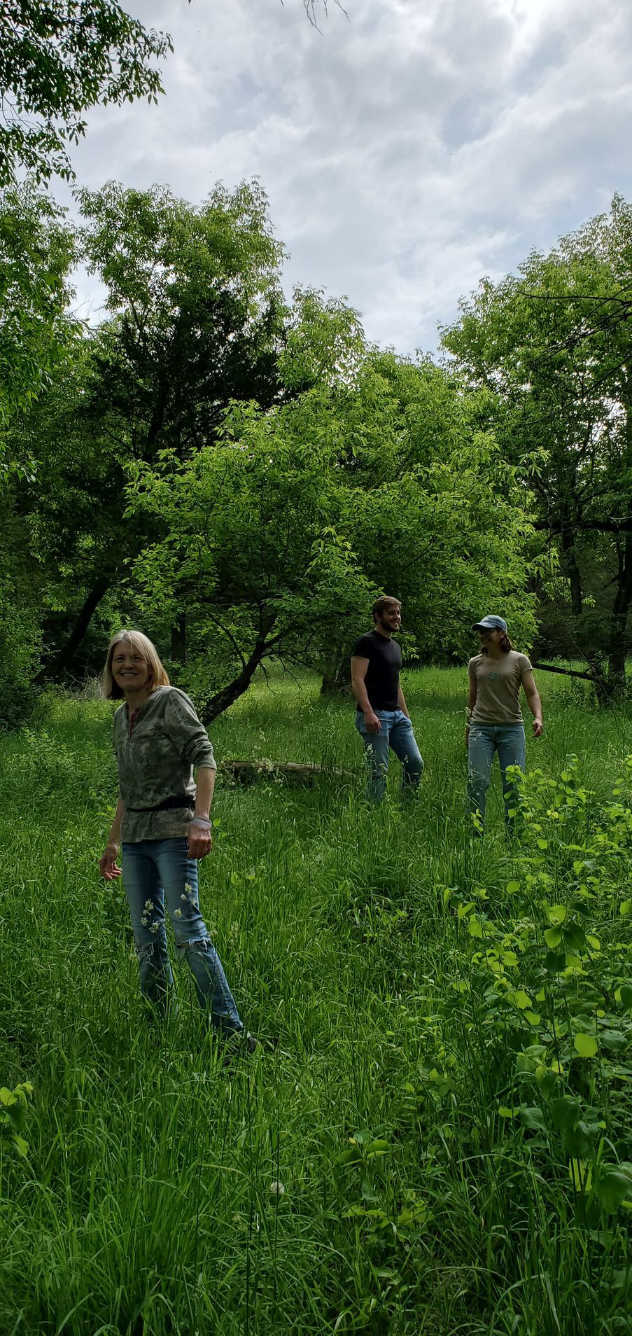 Three people walk through tall green grass in a wooded area under a cloudy sky.