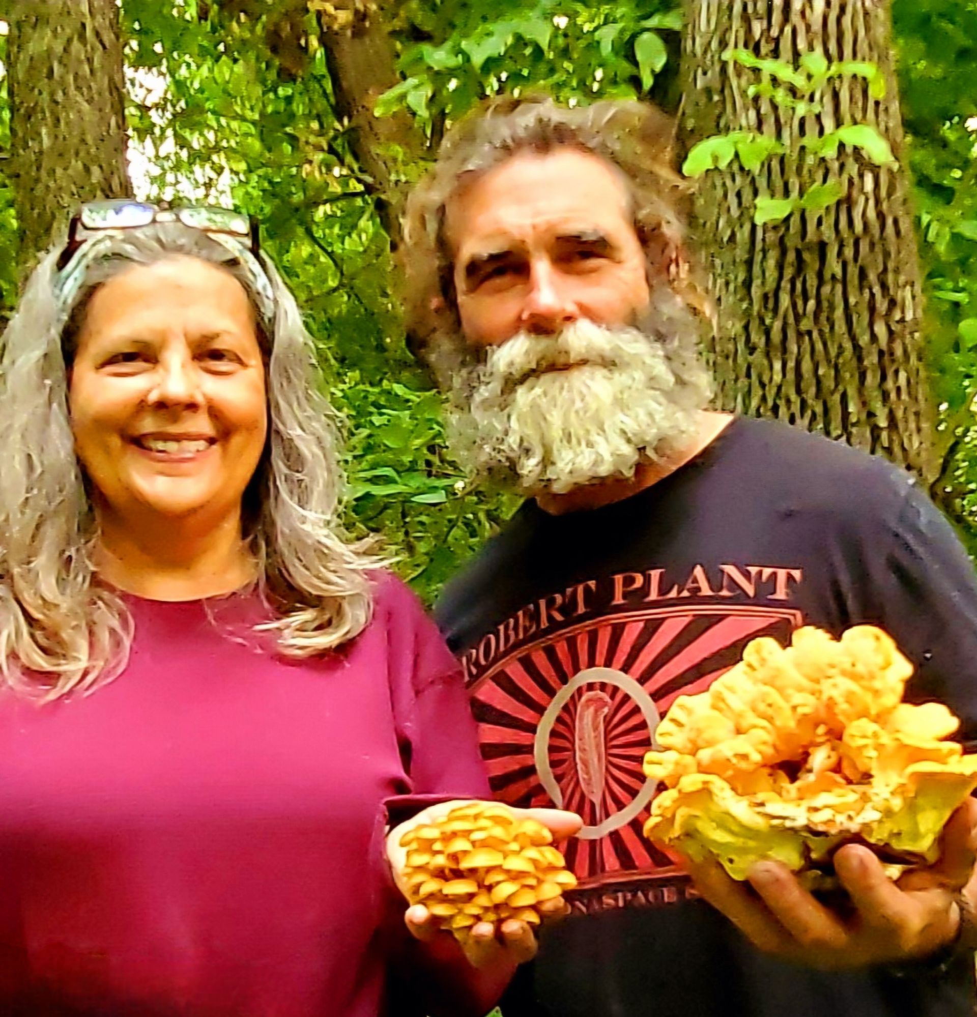 Woman and man holding large yellow mushrooms in a forest, smiling. Man's shirt reads 