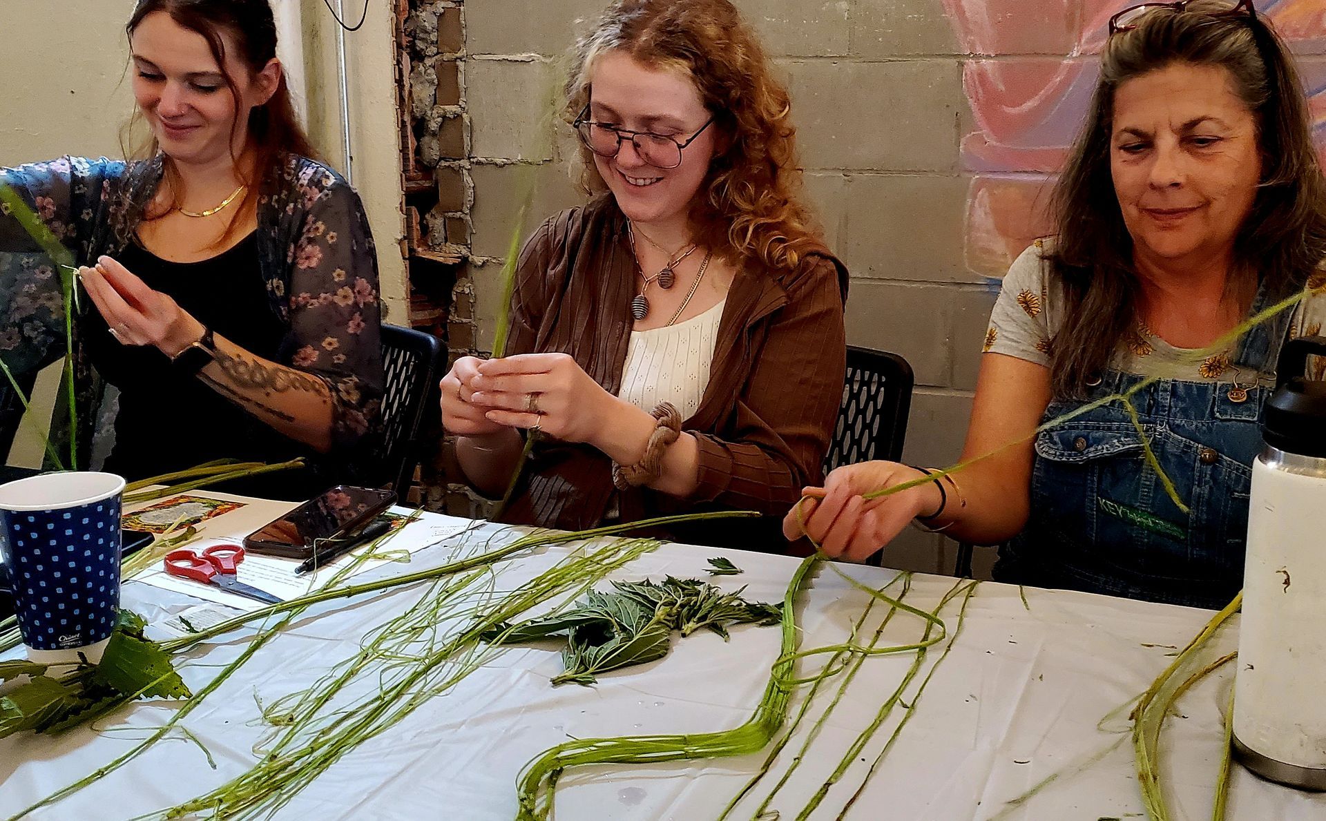 Three people weaving plant fibers at a table, smiling. Indoor setting.