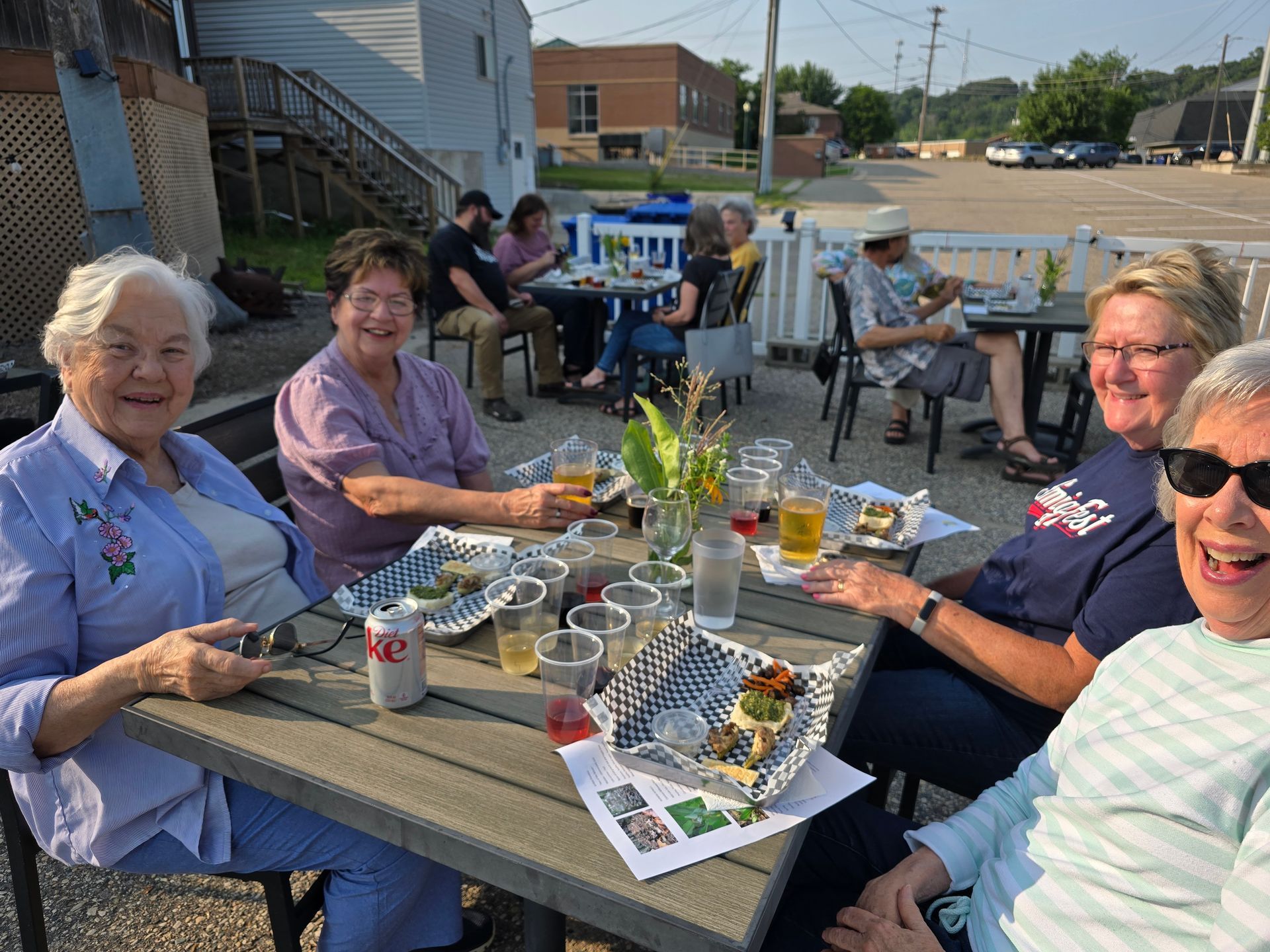 Group of people seated at an outdoor restaurant table. Smiling faces, food, drinks, and a sunny day.