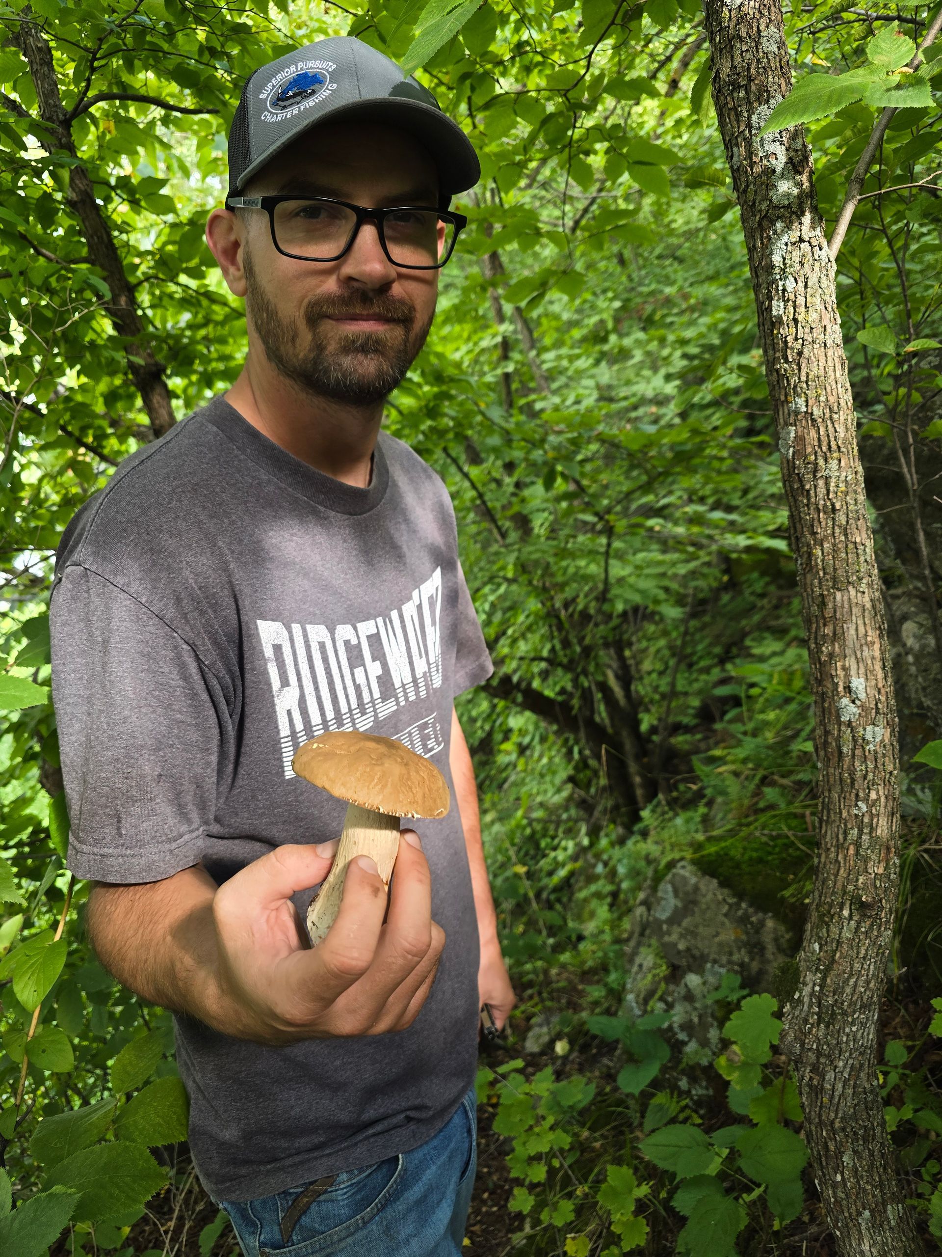 Man in glasses holds a mushroom in a forest setting; he looks toward the viewer.