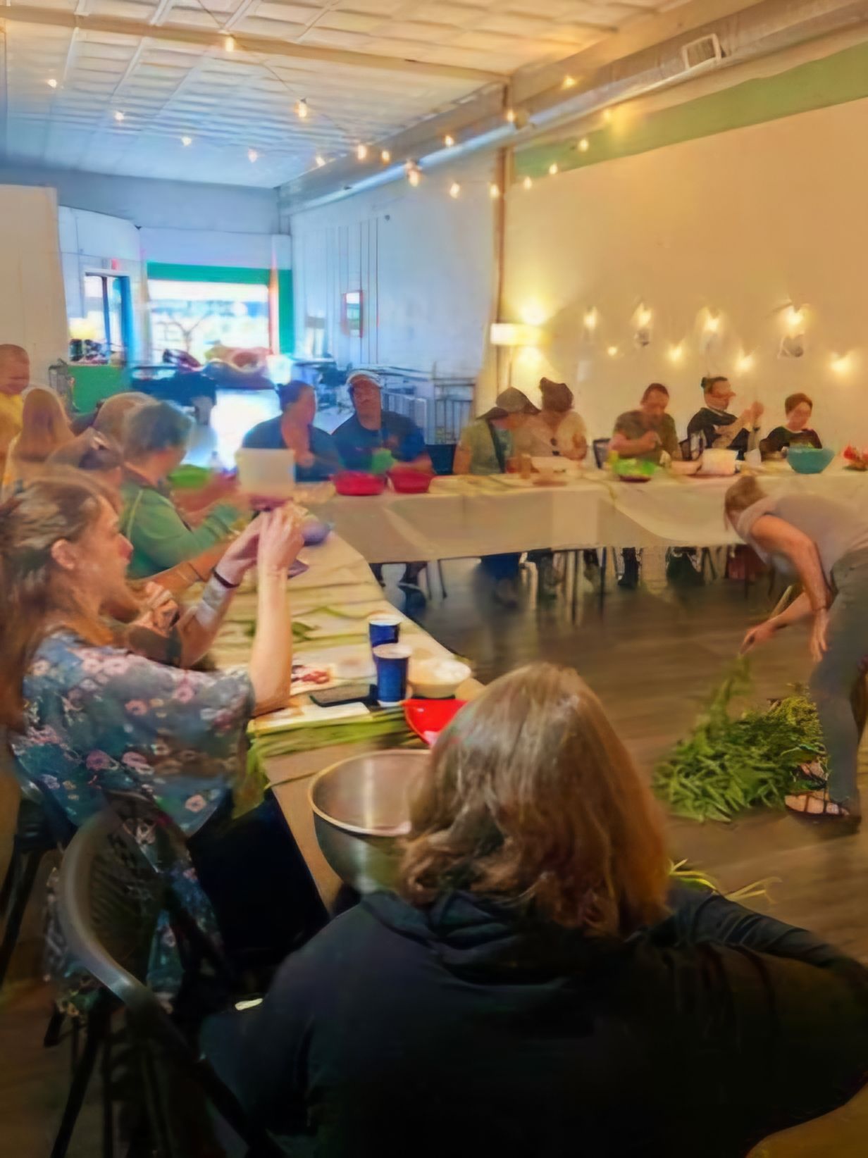 People seated at a table, possibly in a workshop, preparing food with a person nearby handling herbs.