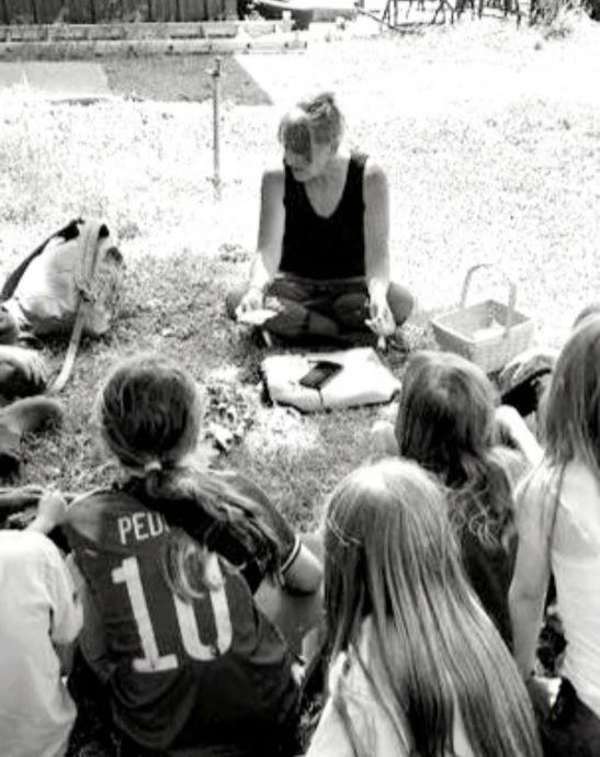 Woman reading to a group of children seated outdoors. White basket nearby.