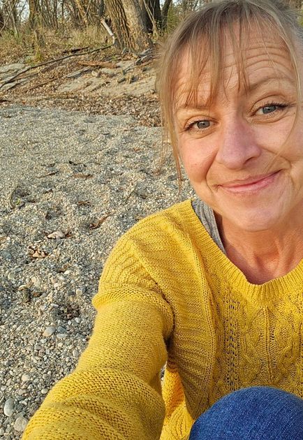 Woman in yellow sweater smiles, sitting on a rocky beach.