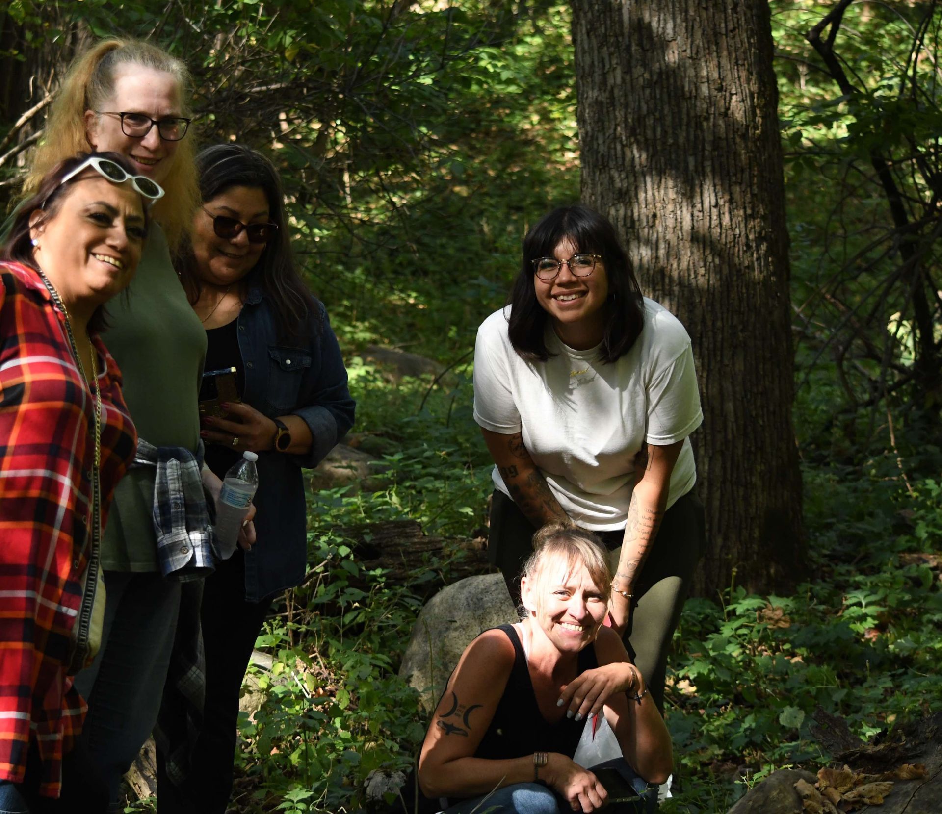 Five people smile in a sunlit forest. One person kneels. Lush green foliage and a tree trunk are visible.