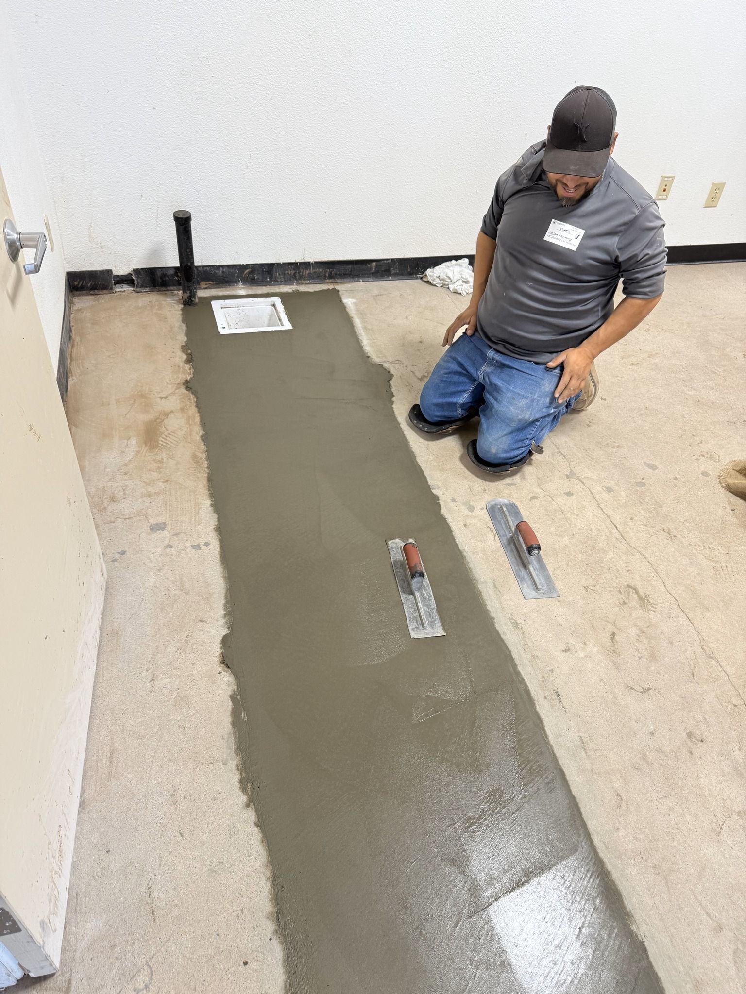 A worker kneeling on a floor, smoothing a strip of wet concrete with a trowel in an indoor space.