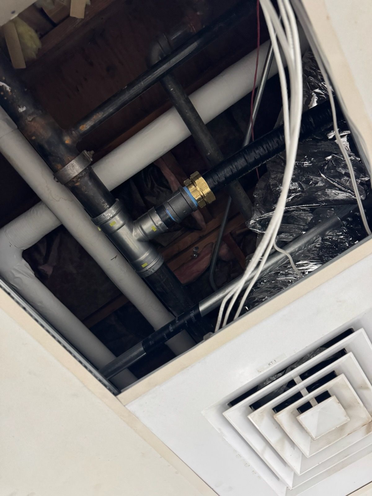 A view inside a ceiling cutout showing various metal and white plastic pipes, wires, and an HVAC air vent.