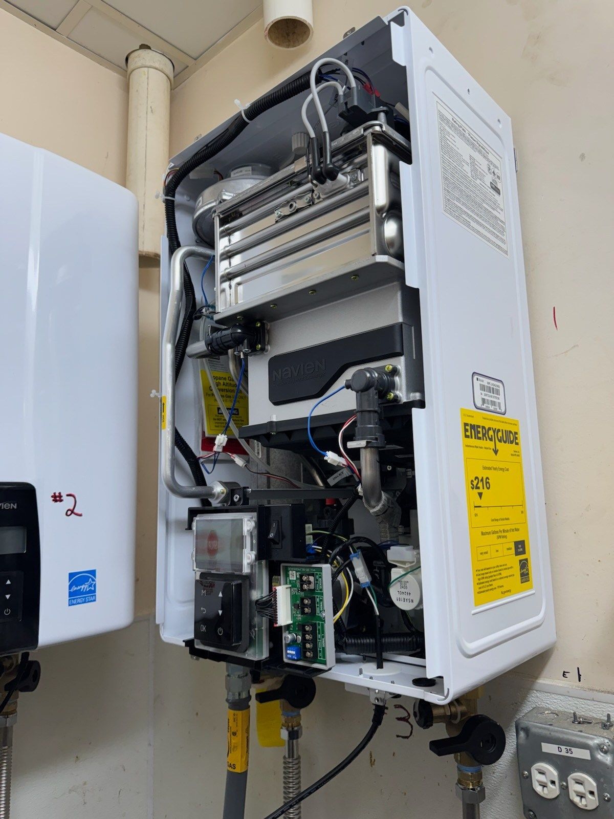 Wall-mounted, open-cased tankless water heater with internal components exposed and a yellow EnergyGuide label attached.