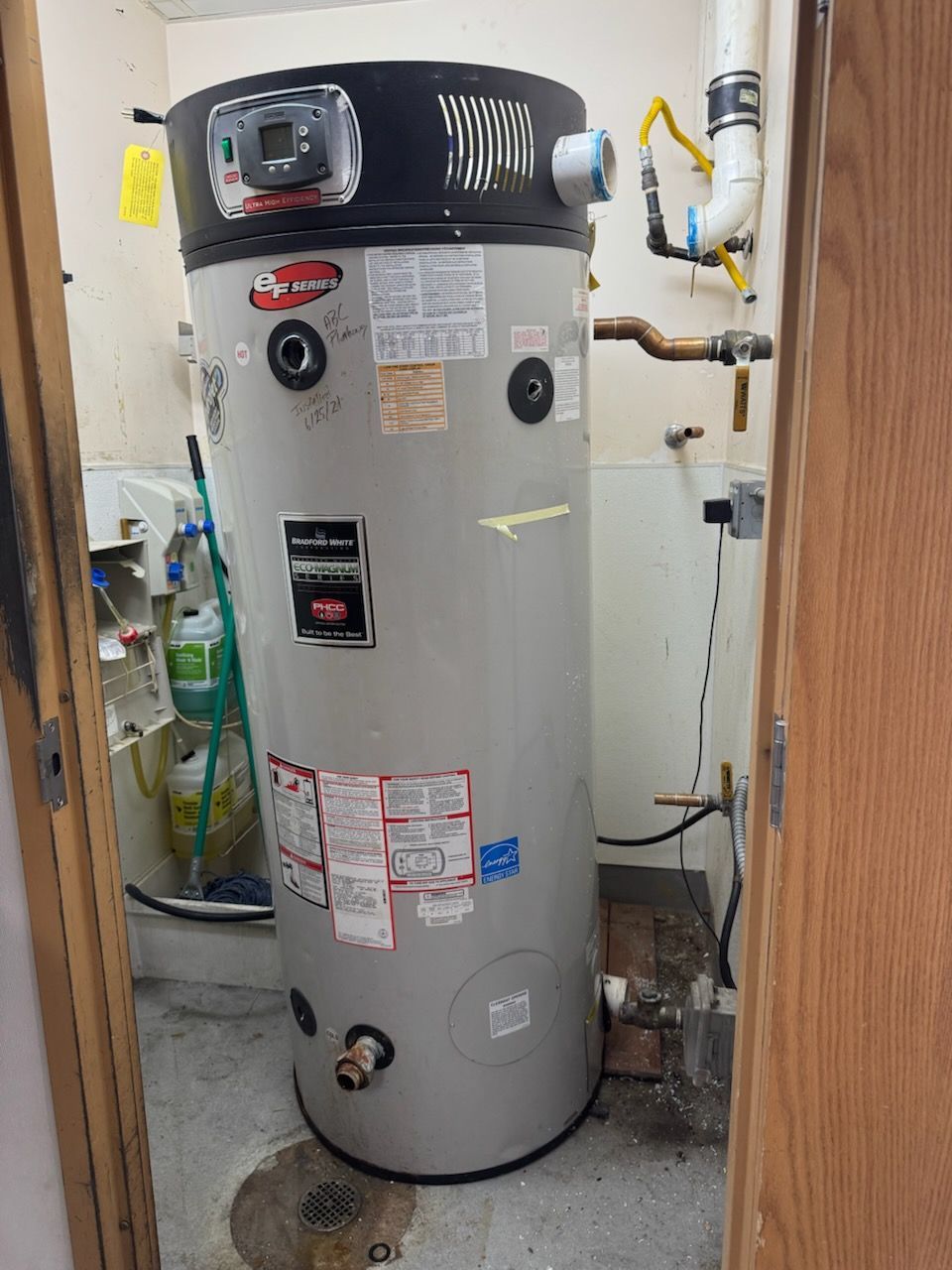 A tall, grey gas water heater stands in a utility closet with connected pipes and a floor drain visible.