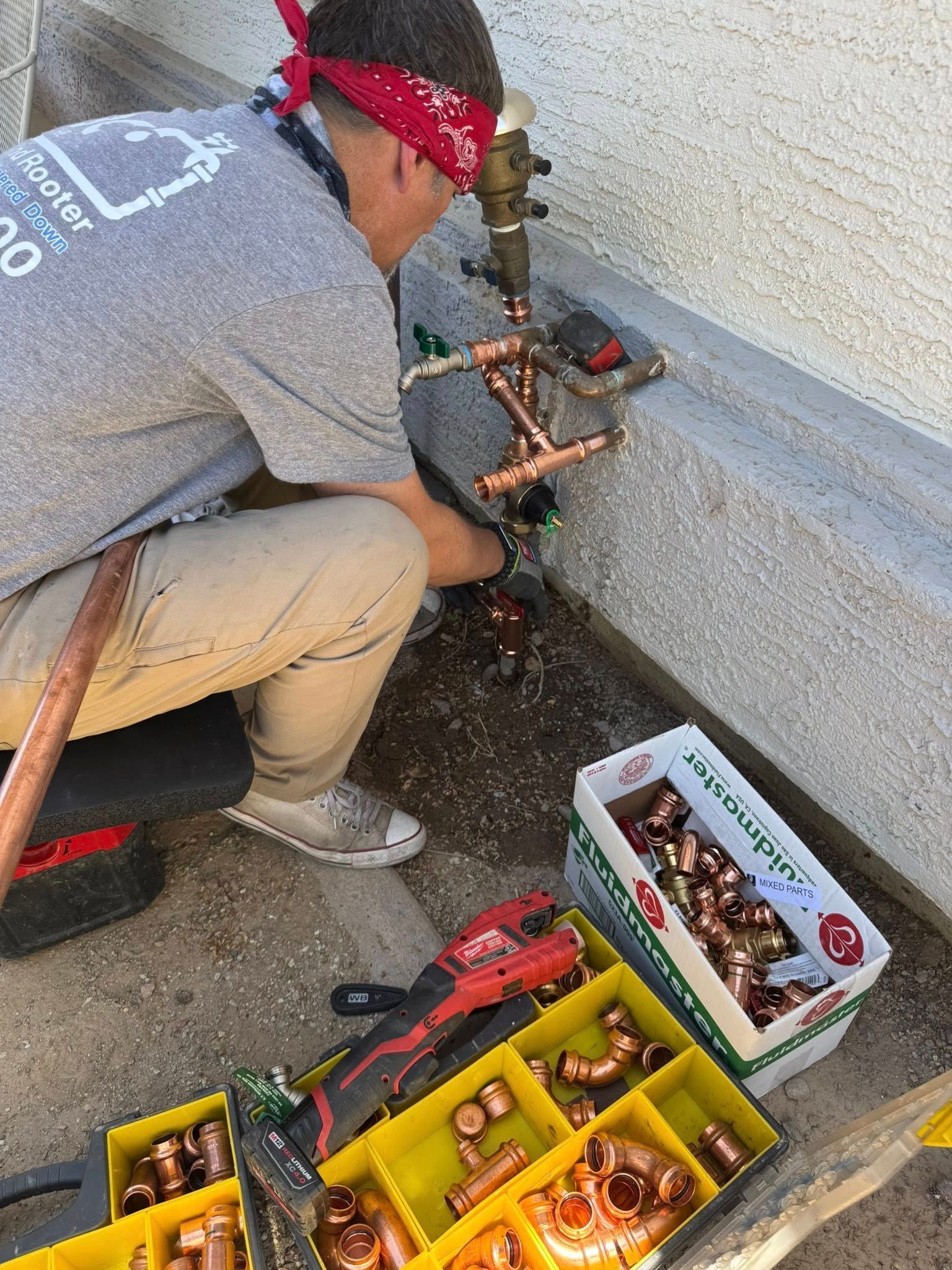 A plumber in a red bandana kneels on concrete, installing copper pipes into an outdoor water valve assembly.