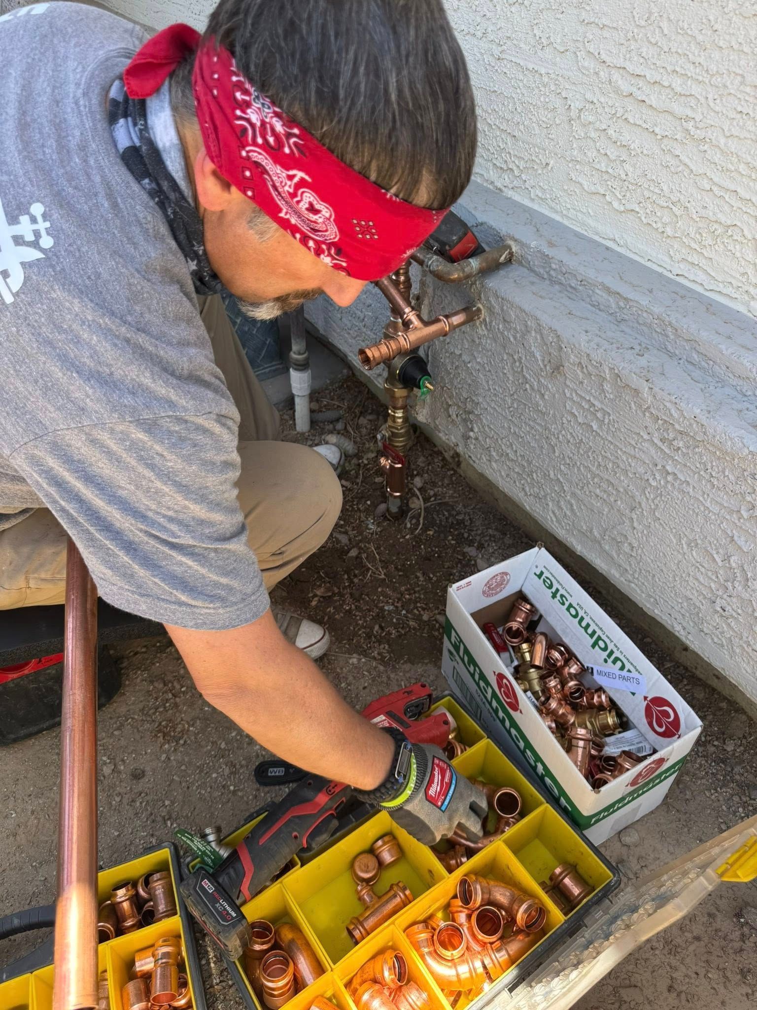 A person wearing a red bandana and gloves repairs copper pipes next to a yellow organizer filled with plumbing fittings.