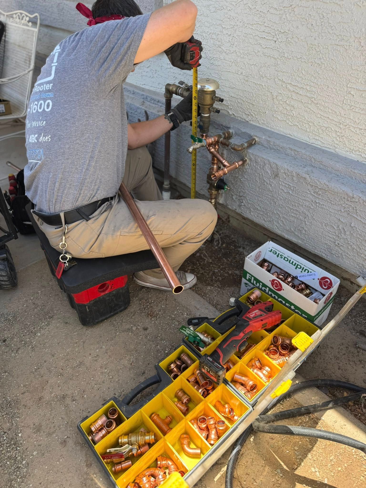 A plumber kneeling outside measures copper piping connected to a home’s water system, with a parts bin nearby.