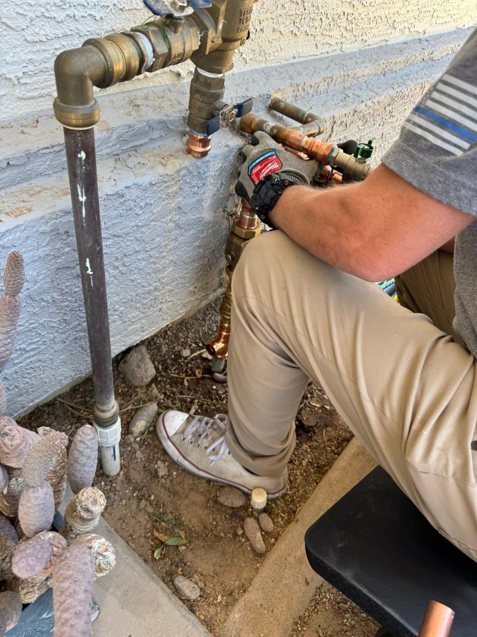 A person kneels by a concrete wall, using tools to repair outdoor plumbing pipes near the ground.