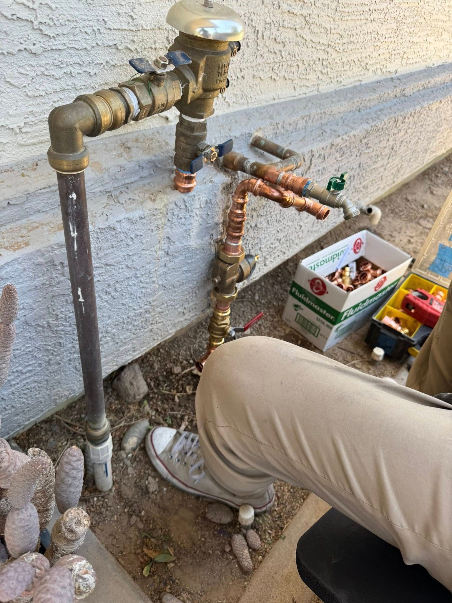 A person in tan pants and white sneakers works on outdoor plumbing with a pressure vacuum breaker and copper pipes.