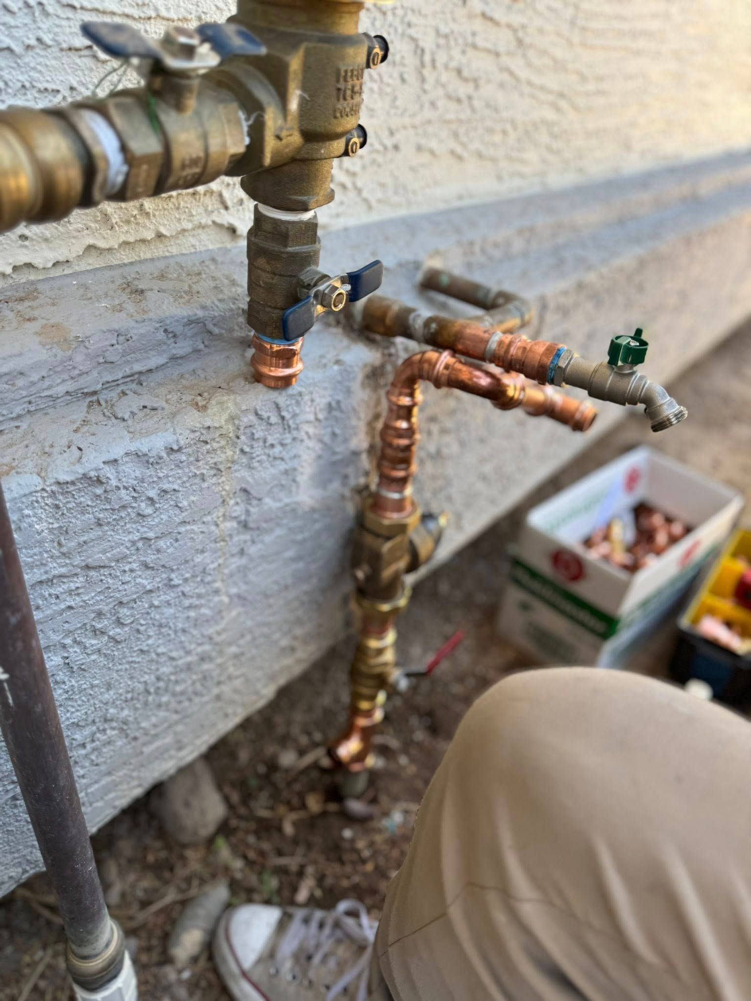 A person performing plumbing repairs on outdoor copper pipes attached to a house foundation.