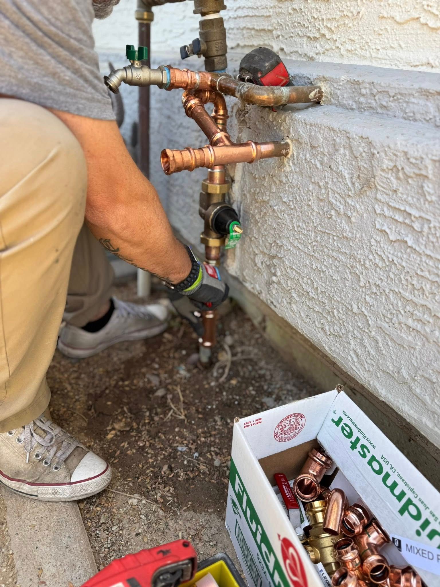 A worker installing copper piping onto an outdoor plumbing assembly near a house wall, with a box of fittings nearby.