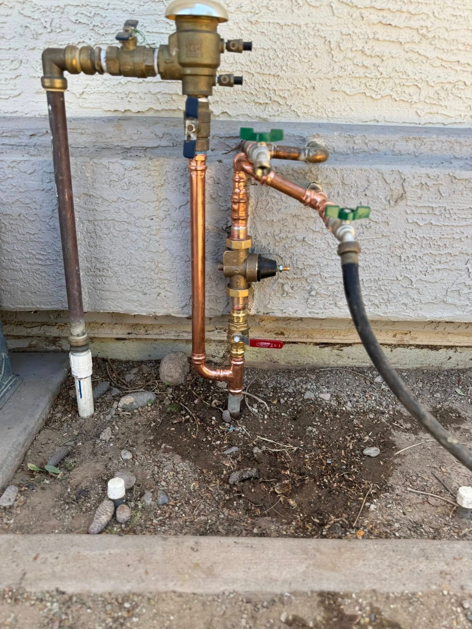 Outdoor irrigation plumbing setup with a brass backflow preventer, copper pipes, valves, and a hose against a wall.