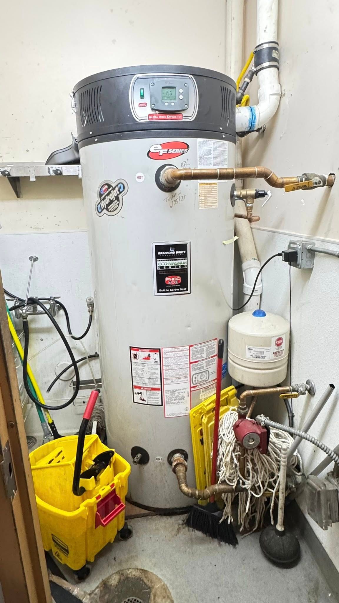 A grey industrial water heater stands in a utility room next to plumbing pipes, an expansion tank, and cleaning supplies.