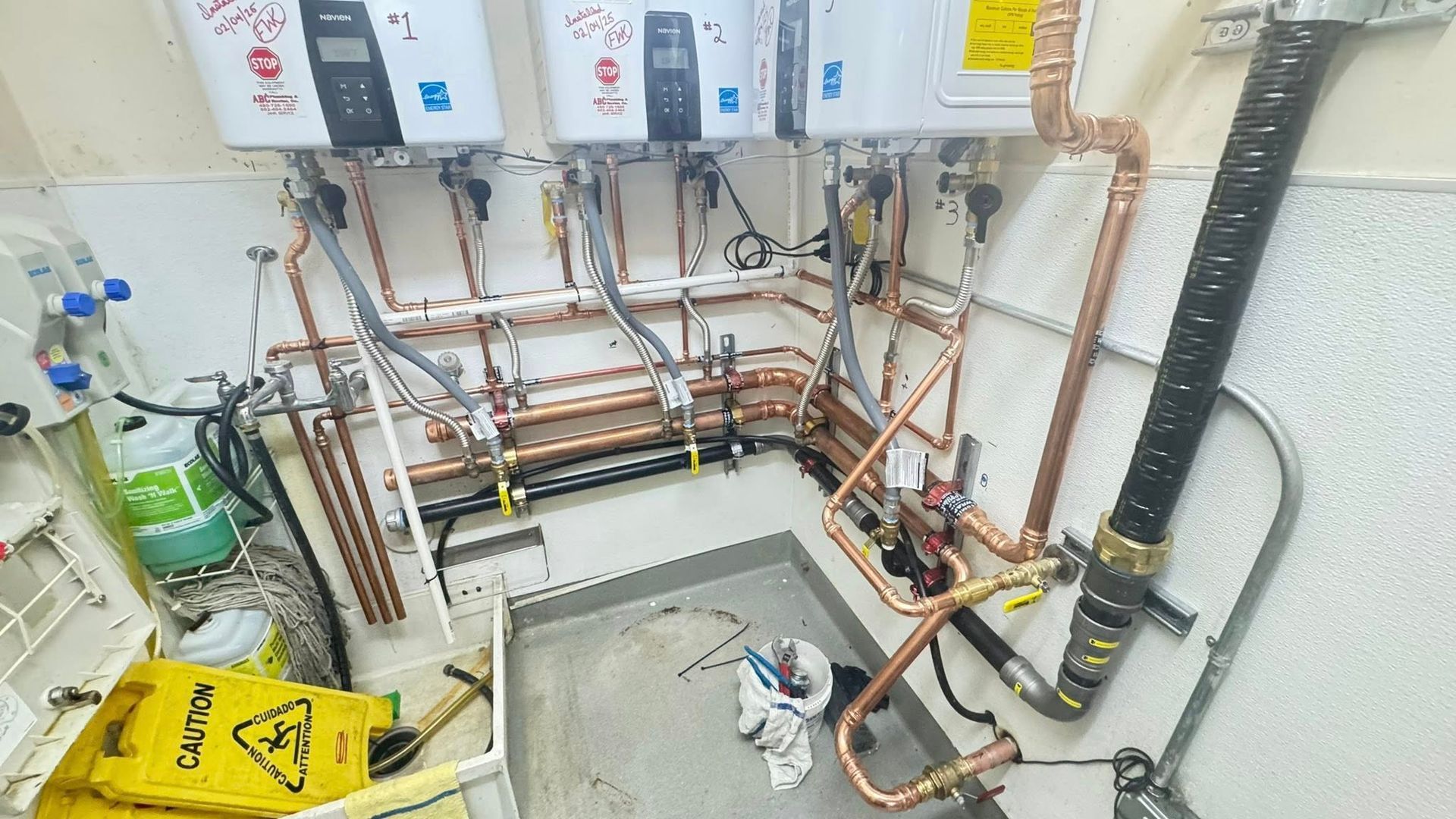 Three commercial water heaters wall-mounted above a copper pipe manifold system in a utility room.