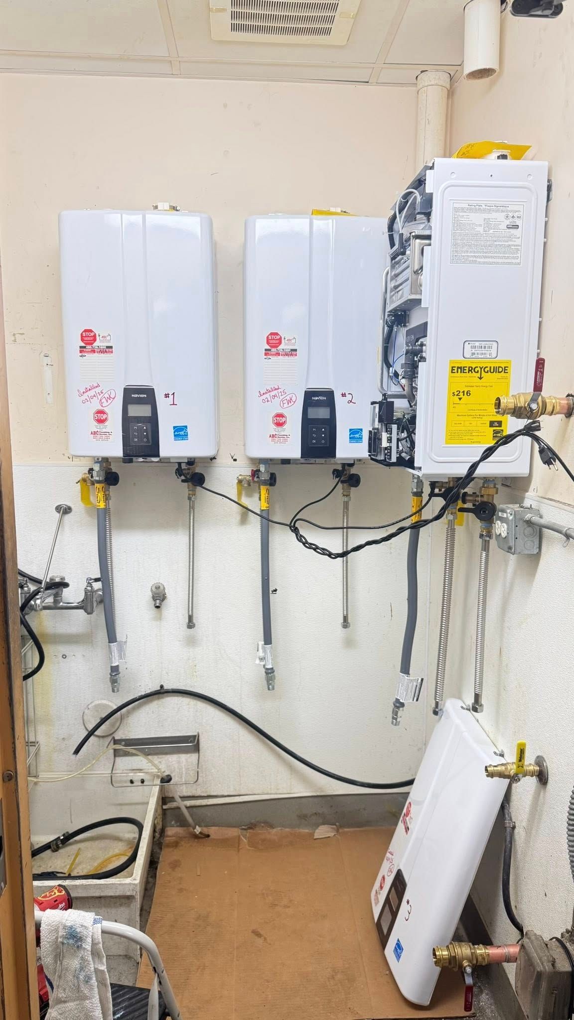 Three white tankless water heaters mounted on a wall, with another unit sitting on the floor in a utility room.