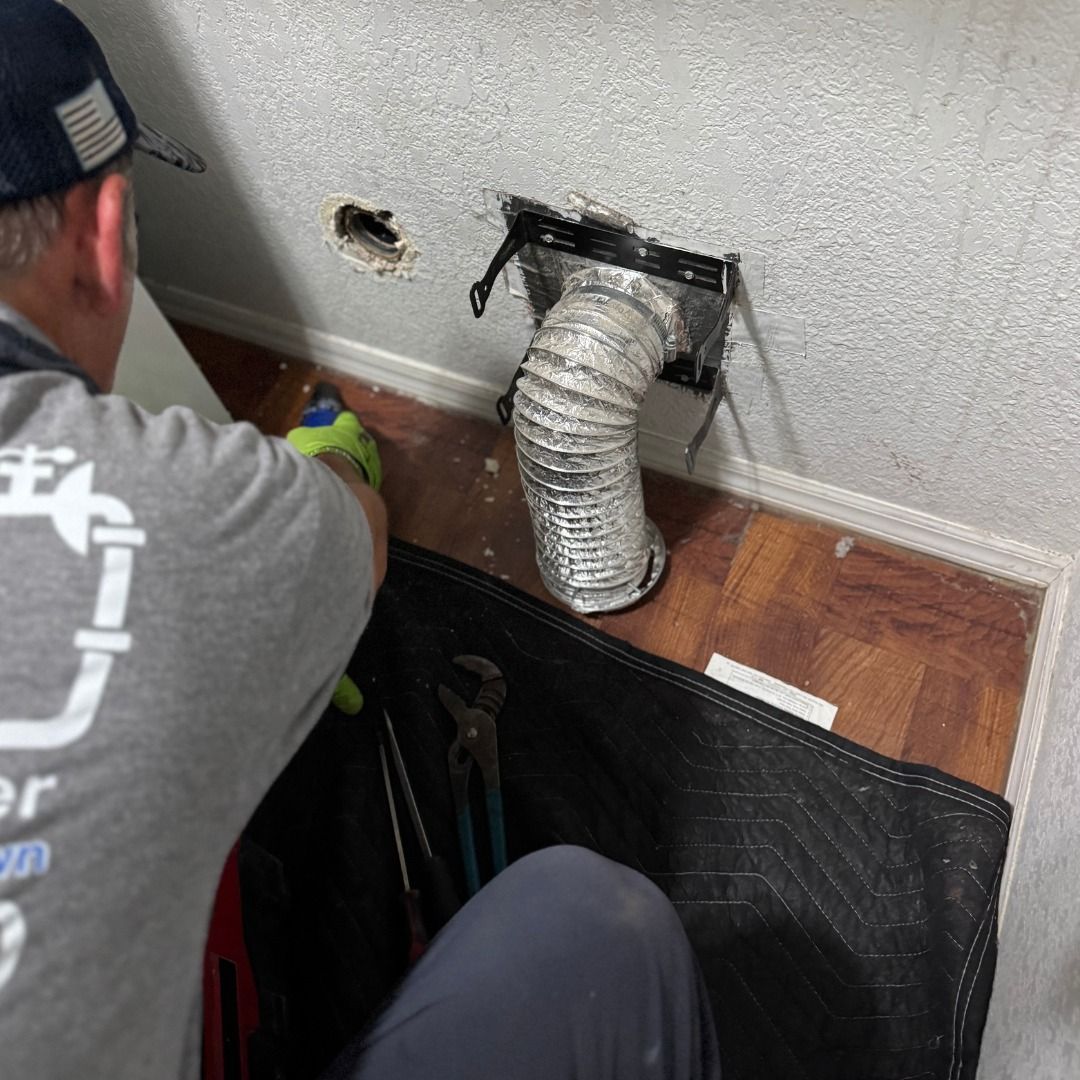 A technician in a gray shirt works on a wall-mounted dryer vent hose in a room with wood-look flooring.