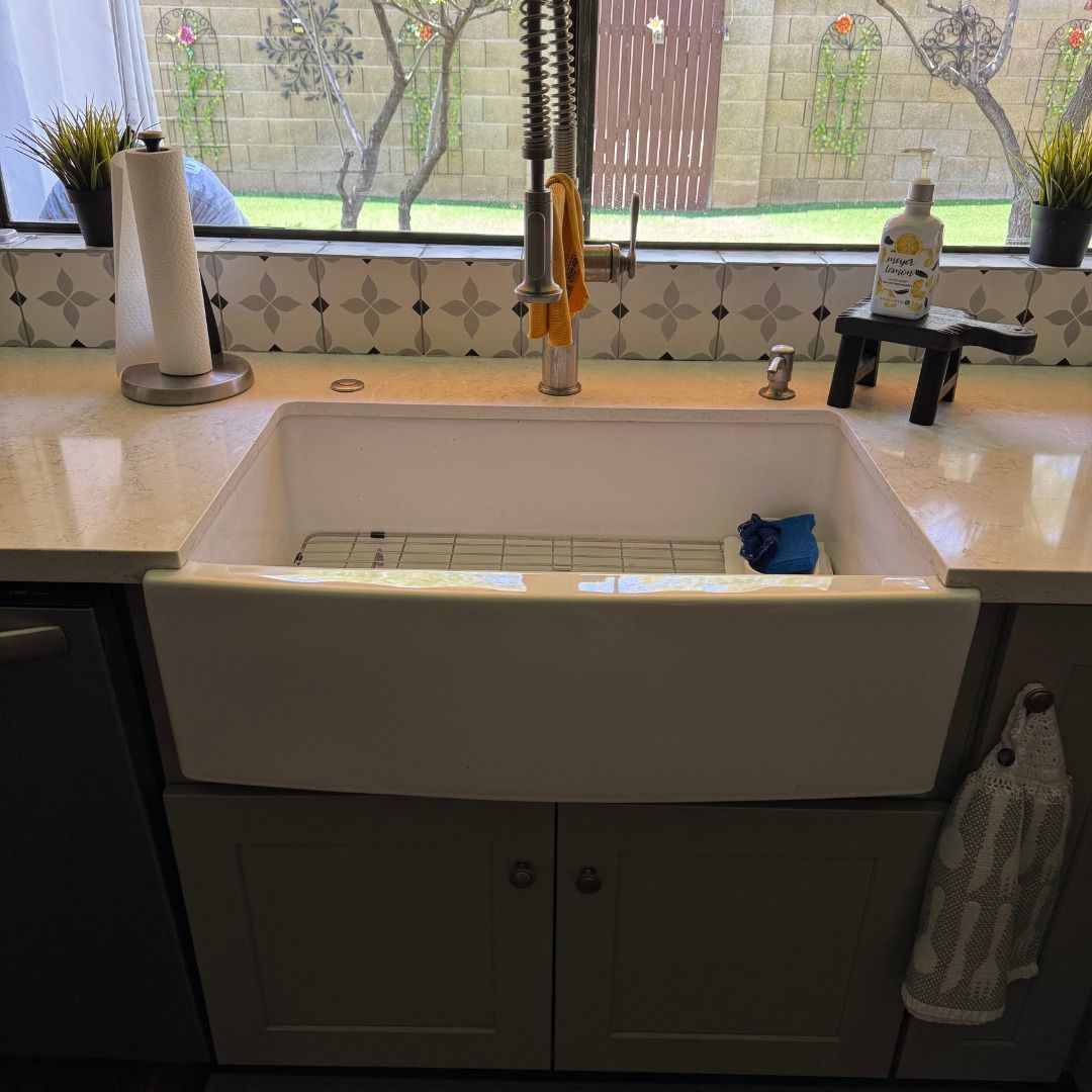 White farmhouse sink with a wire rack and sponge in a kitchen with gray cabinets and a patterned tile backsplash.