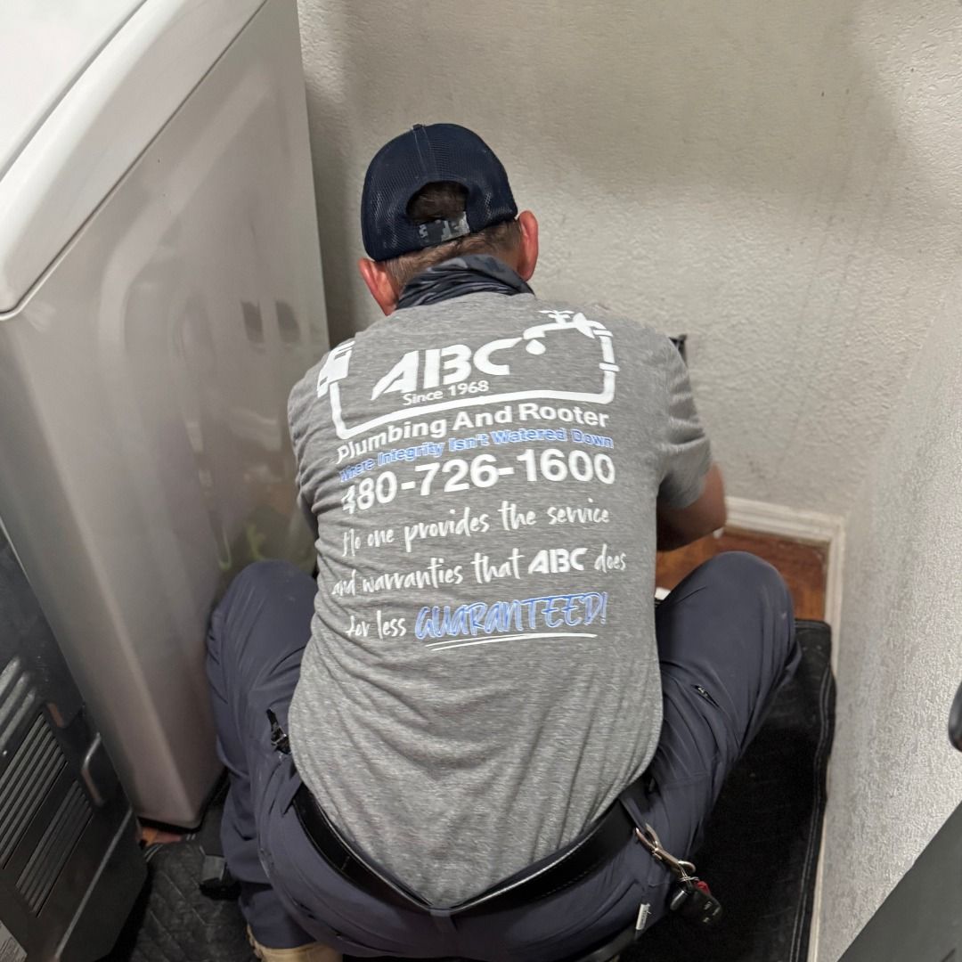 A plumber in a grey company uniform crouches on a floor mat while working behind a white laundry appliance.