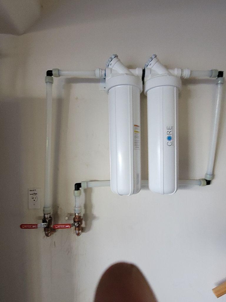 Two vertical white water filter canisters mounted on a wall with connected white plastic piping and two shut-off valves.