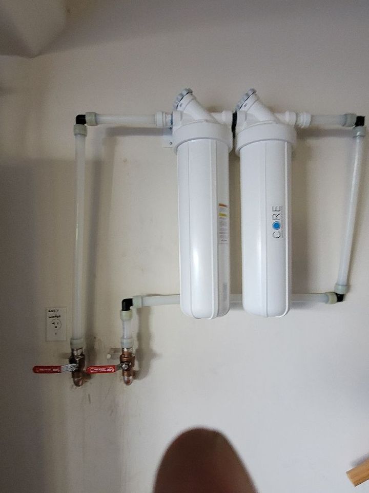 Two vertical white water filter canisters mounted on a wall with connected white plastic piping and two shut-off valves.