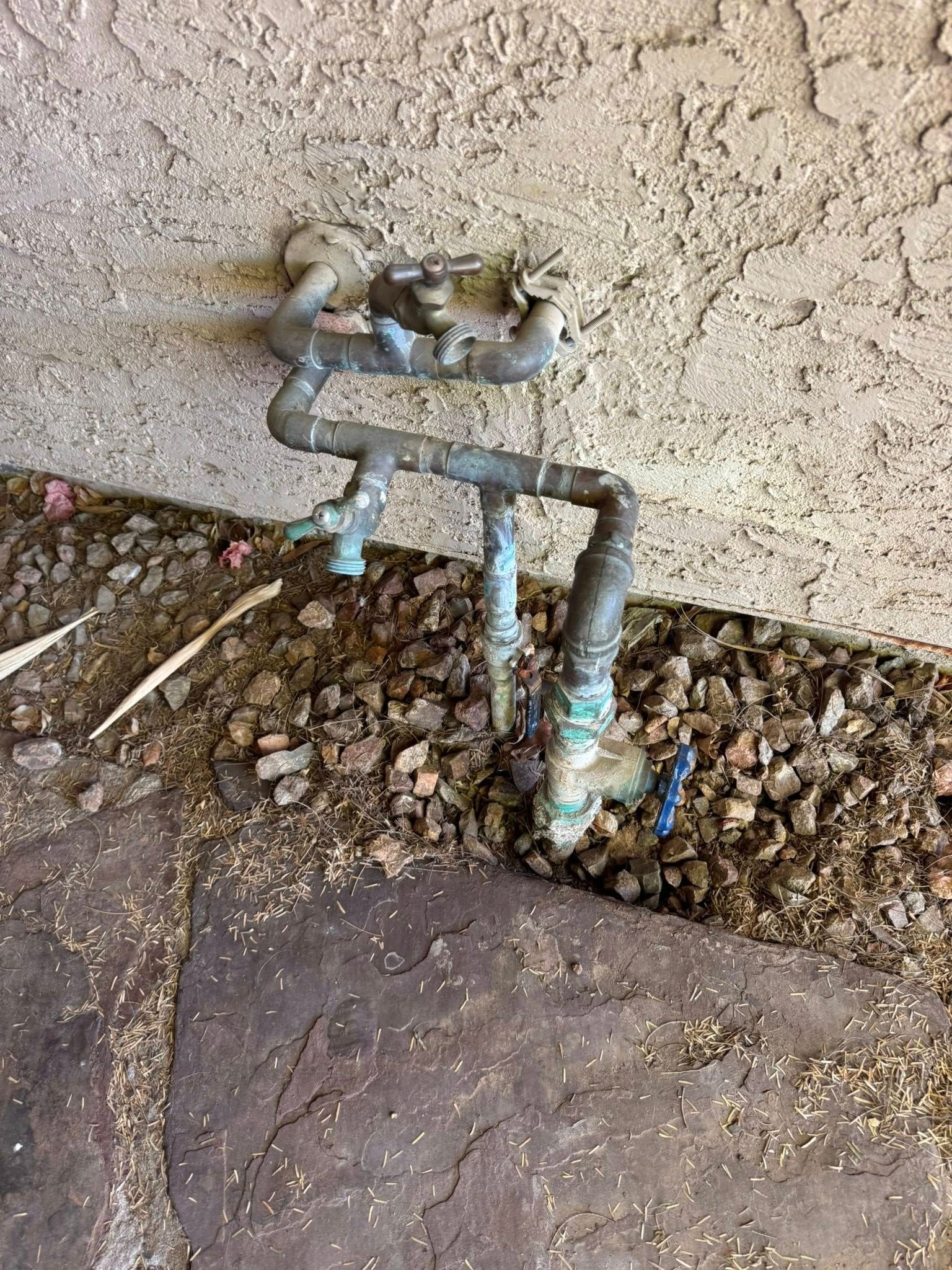 Copper plumbing pipes with multiple valves and a shut-off handle emerging from the ground against a stucco wall.