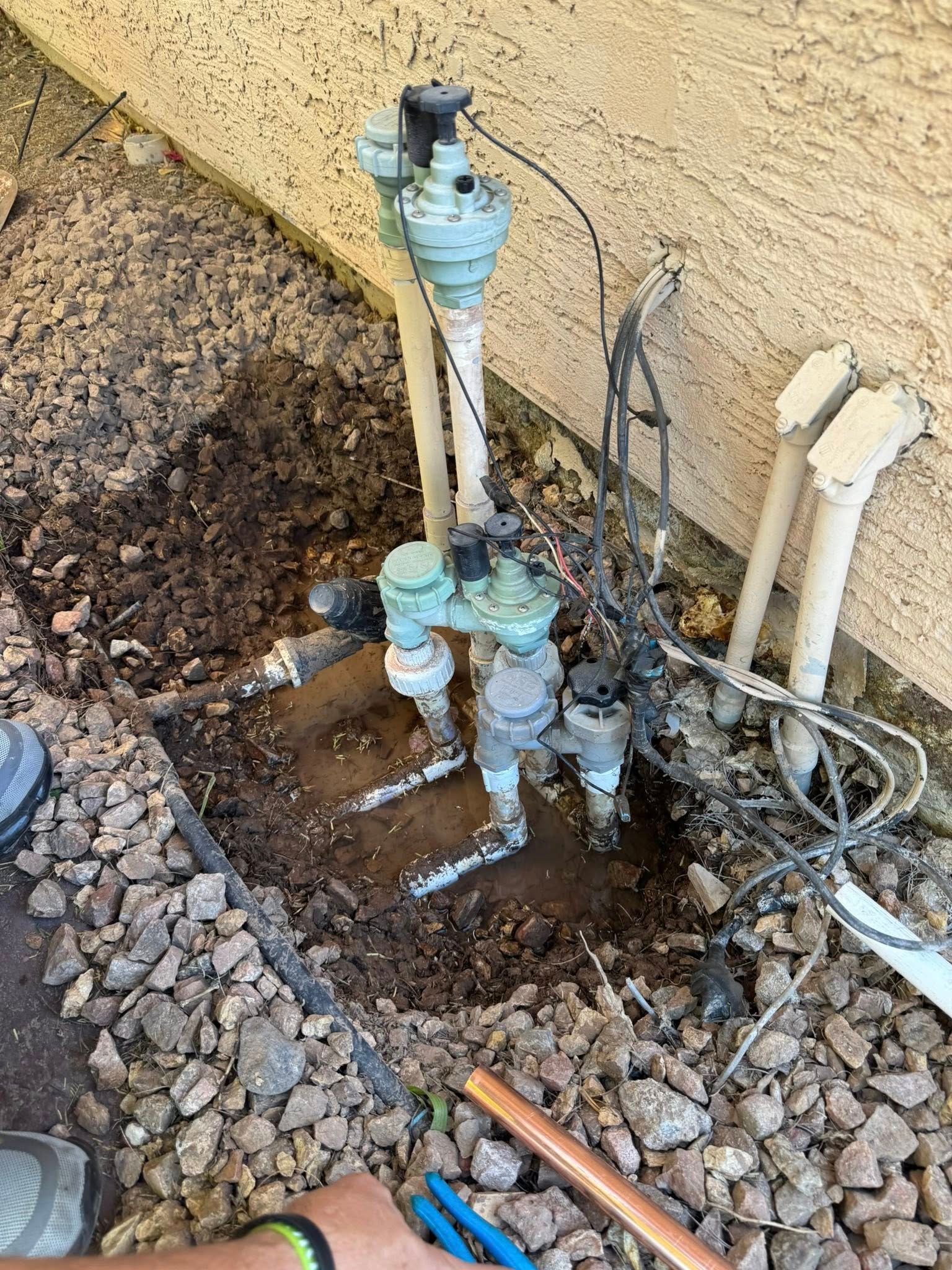 Outdoor irrigation valve manifold assembly partially exposed in a dug-out trench next to a tan stucco wall.