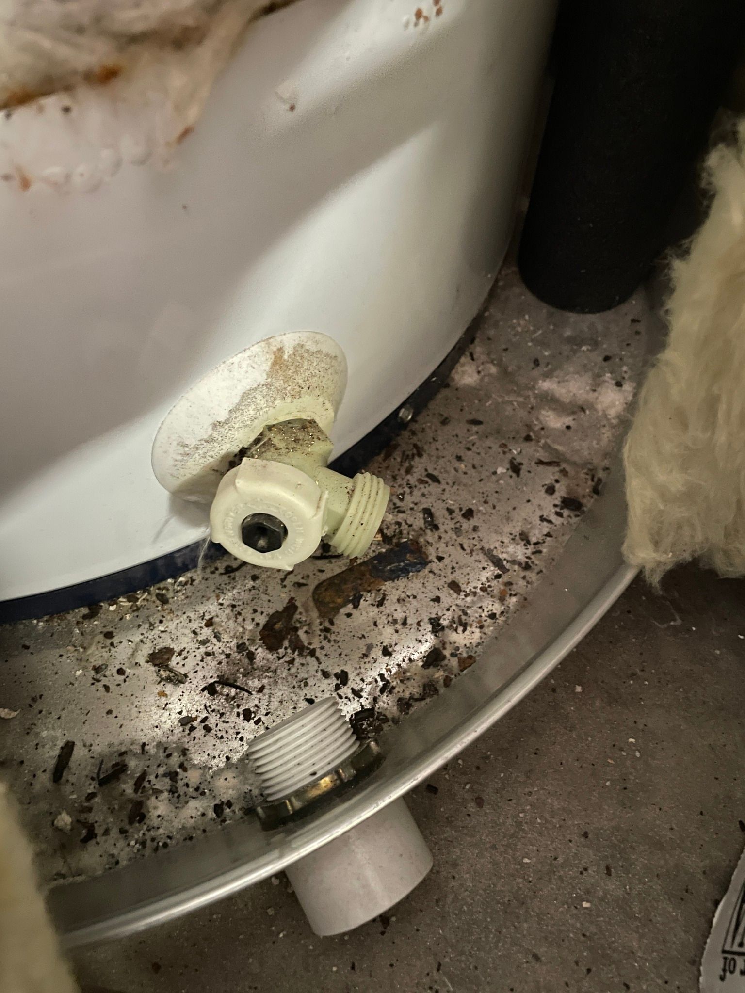 A close-up of a white plastic water heater drain valve and a threaded metal pipe outlet in a dirty, debris-filled area.