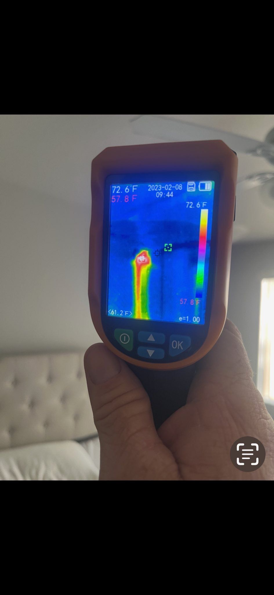 A hand holds a thermal imaging camera, displaying a heat signature on a wall in a bedroom.