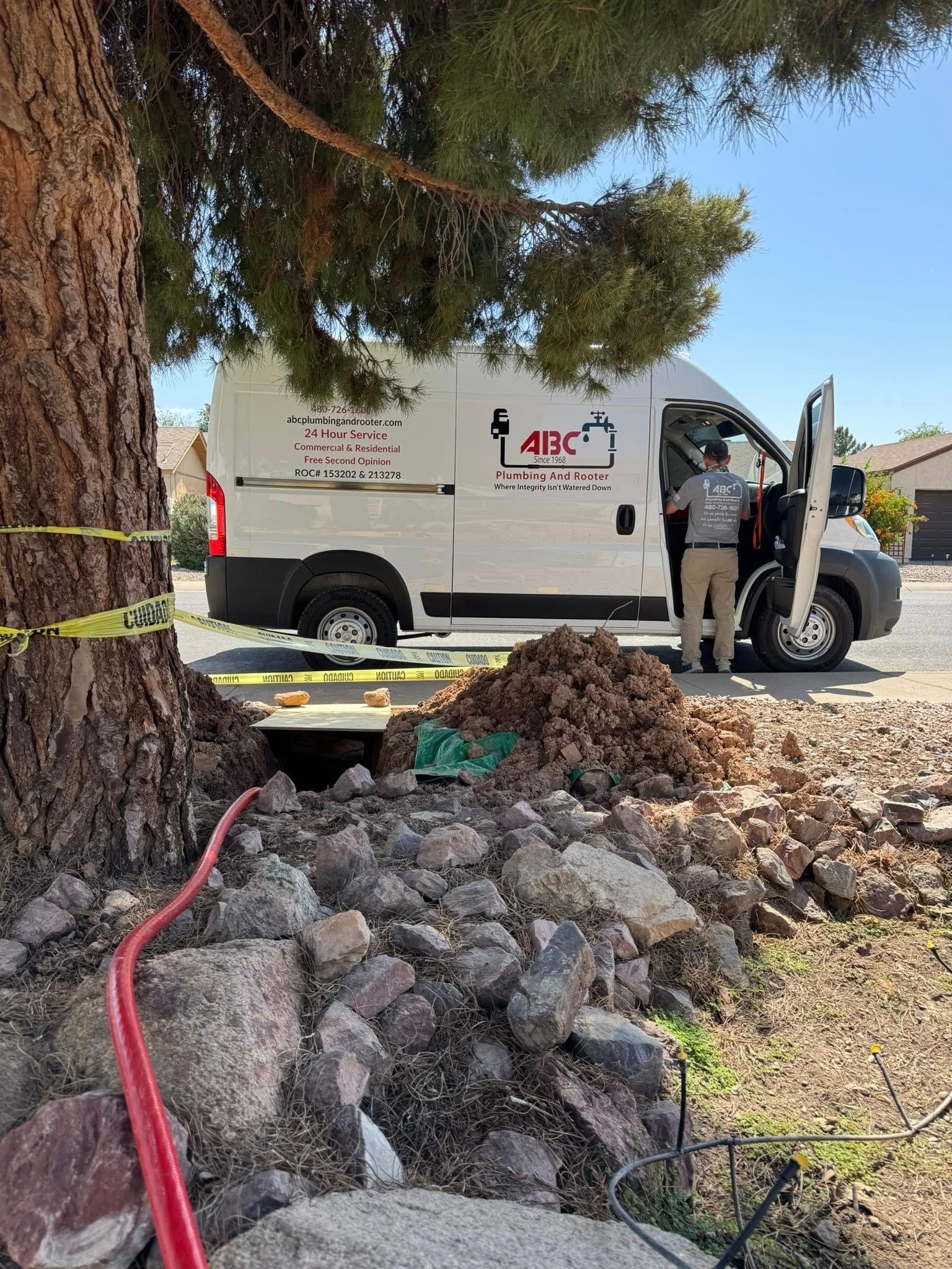 A technician stands by an open service van near a tree with a yellow tape barrier and excavated dirt on a residential lot.