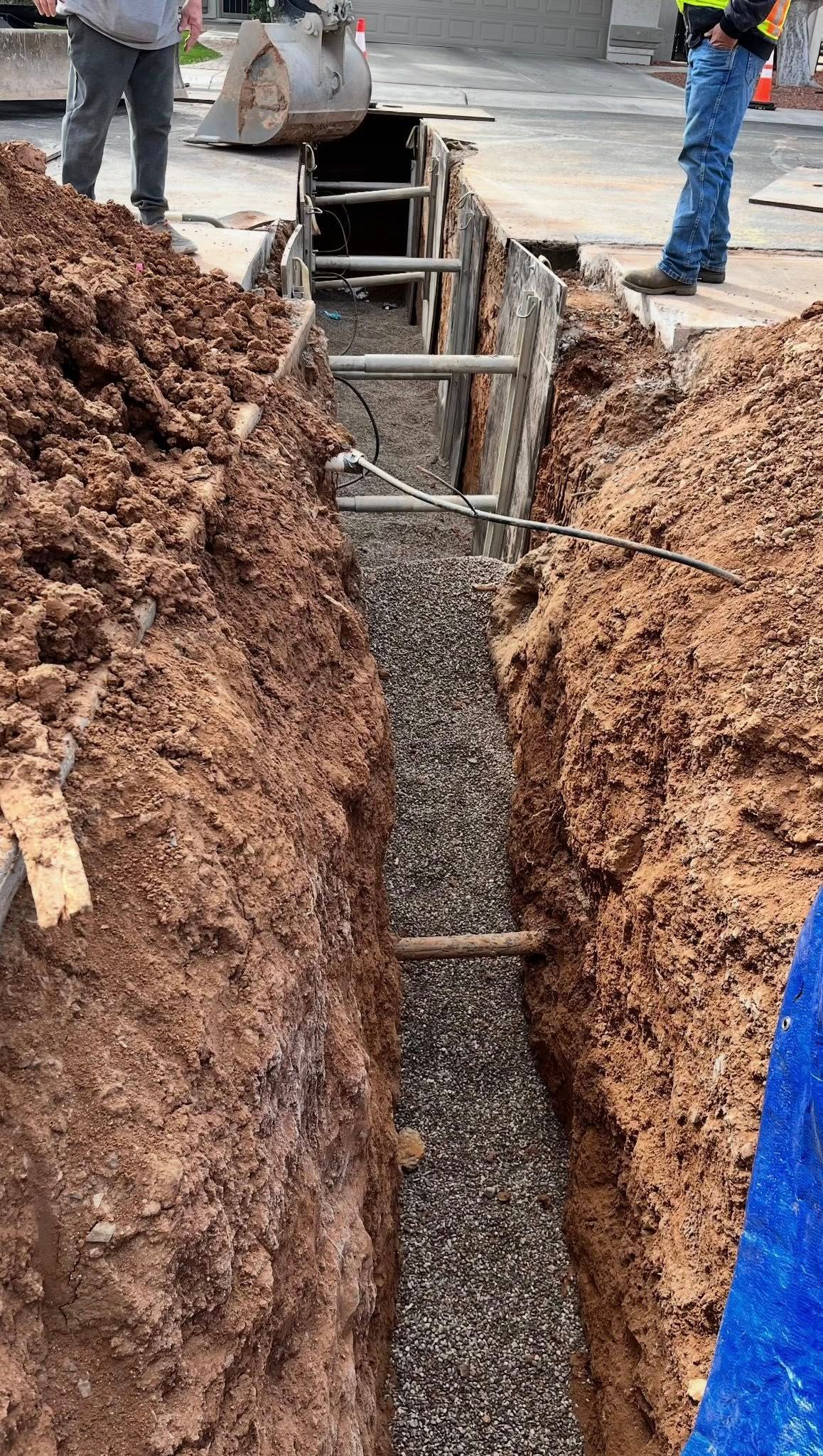 A deep construction trench with shoring, gravel at the bottom, and workers standing on the edge.