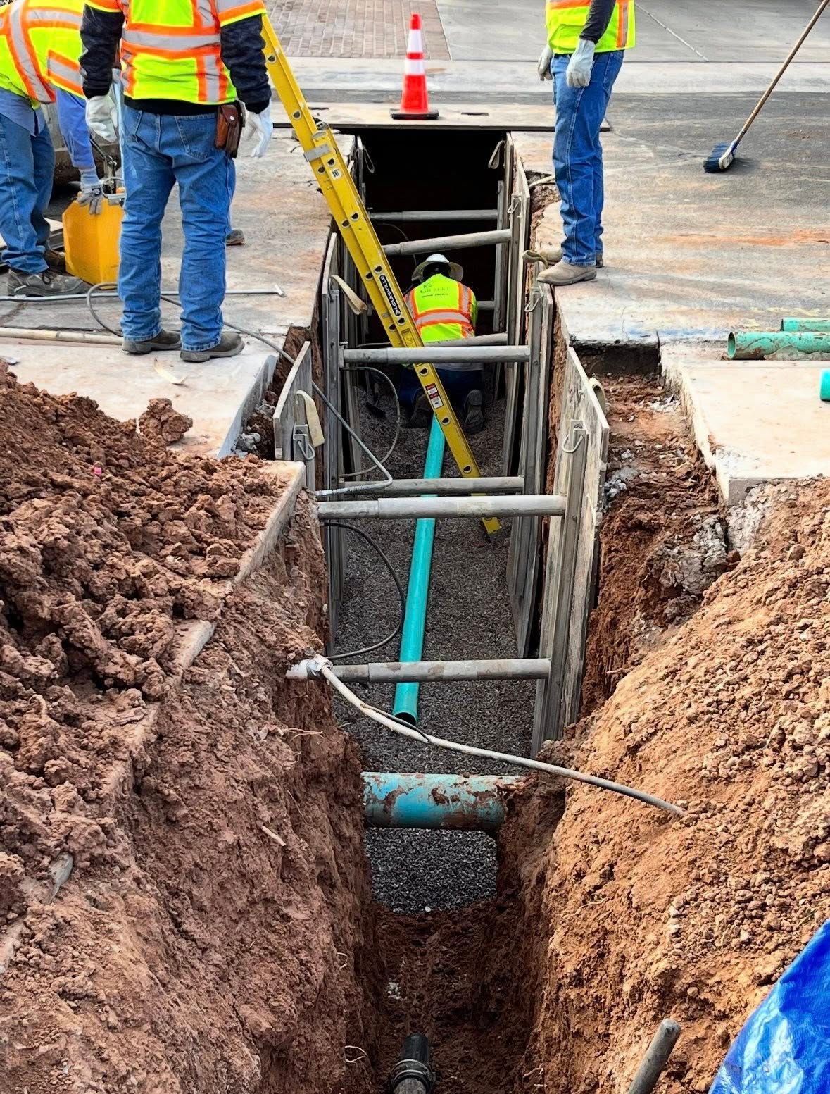 Construction workers in high-visibility vests work in a narrow, shored-up trench with a blue pipe and gravel bedding.