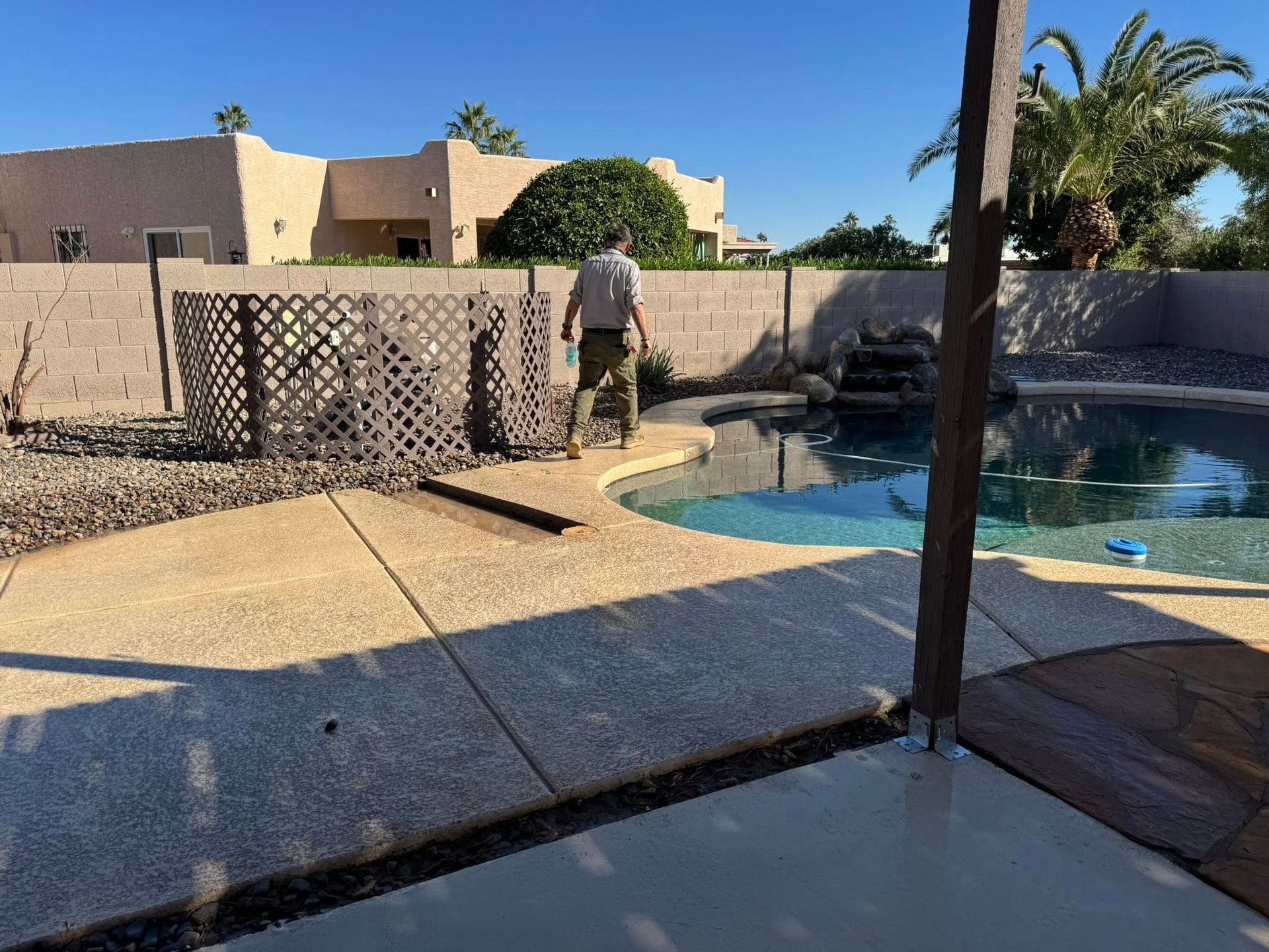 A person in tan clothes walks near a swimming pool in a desert-style backyard with a beige house and lattice fence.
