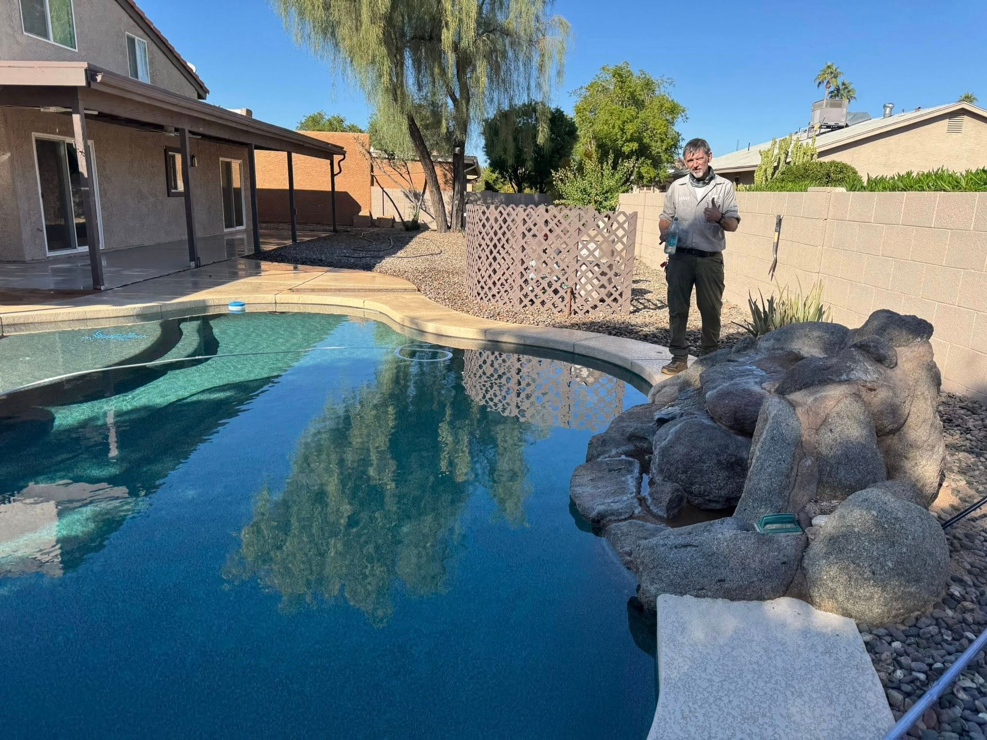A person stands by a backyard pool with a rock water feature, a house, and desert landscaping in the background.