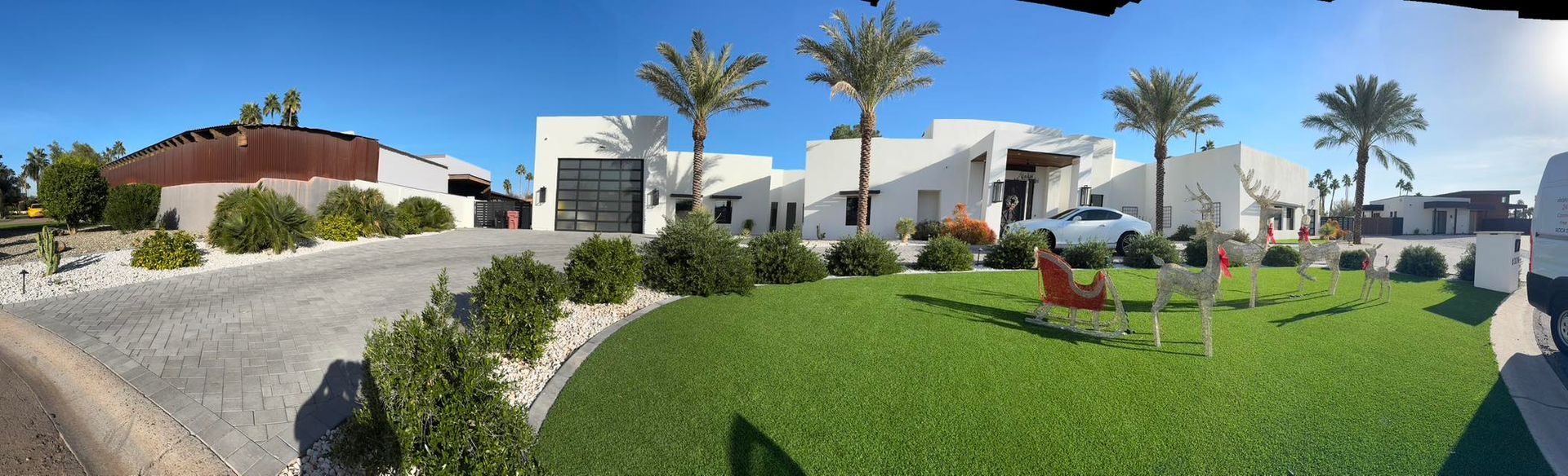 A panoramic view of a white, modern house with a green lawn, palm trees, and a parked car under a clear blue sky.