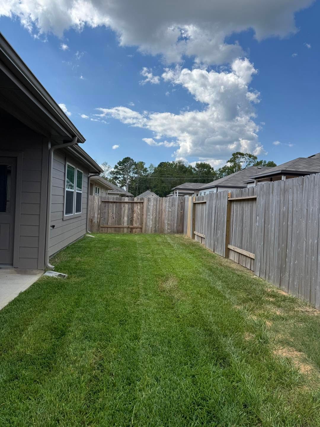 Narrow grassy side yard between two houses, with wooden fences and a partly cloudy blue sky.