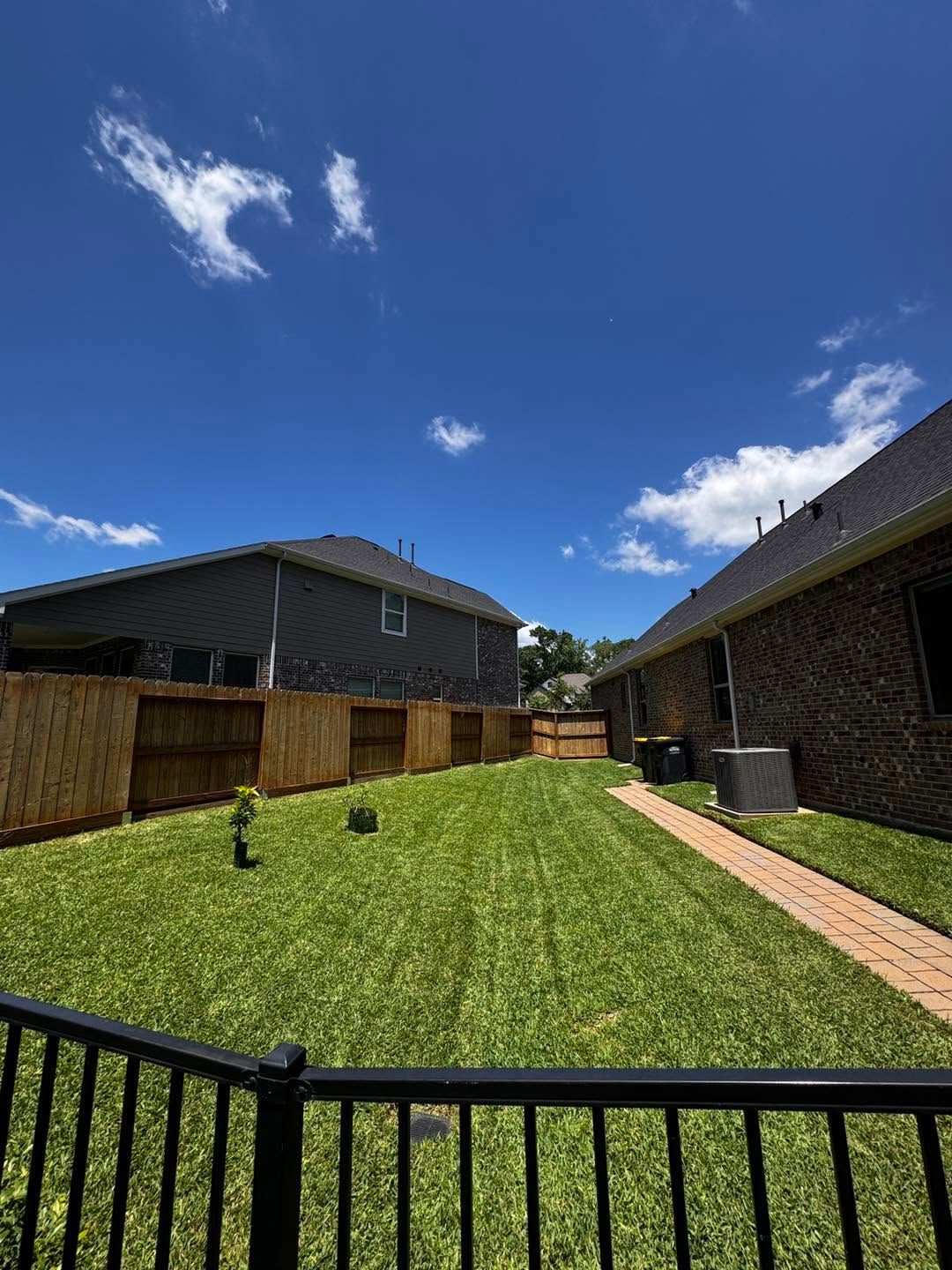 Backyard with green lawn, wooden fence, brick houses, and blue sky with clouds